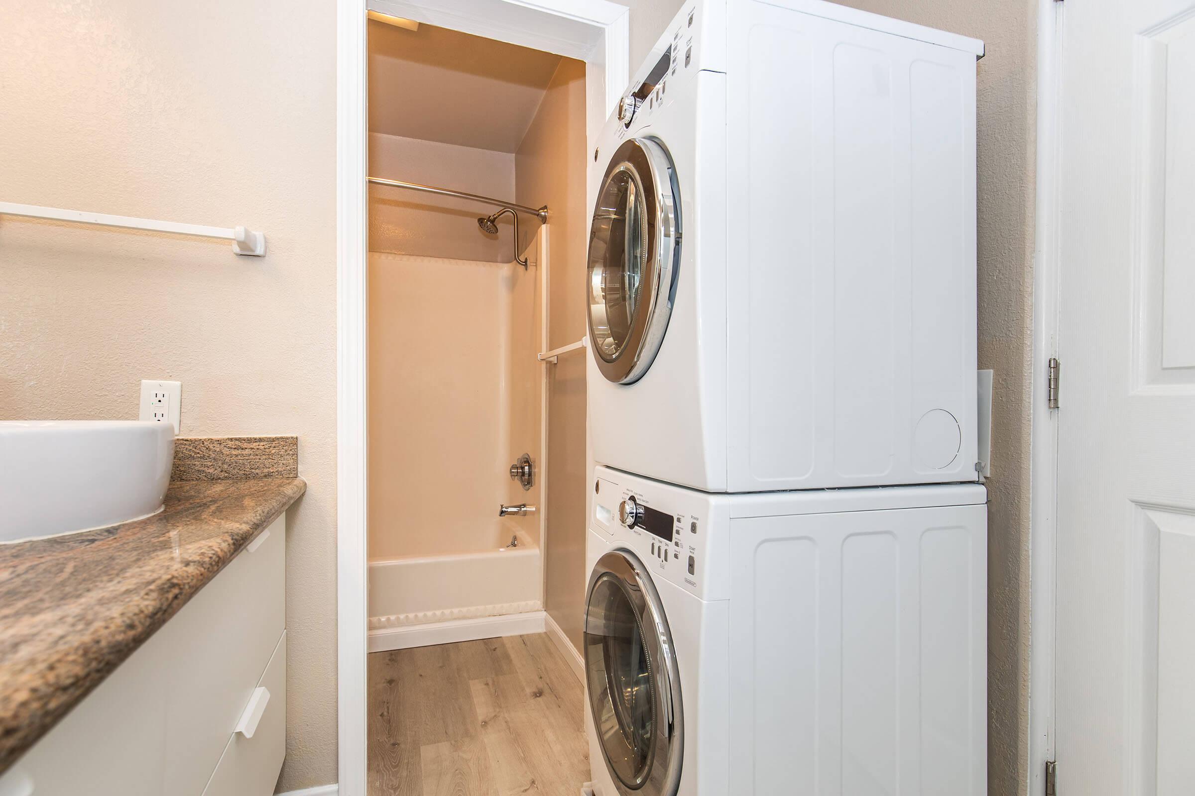 A compact laundry room featuring a stacked washer and dryer in white, positioned next to a sink with a granite countertop. A shower area is visible in the background, with light-colored walls and a simple design. The floor has a wood-like finish, contributing to a modern and functional space.
