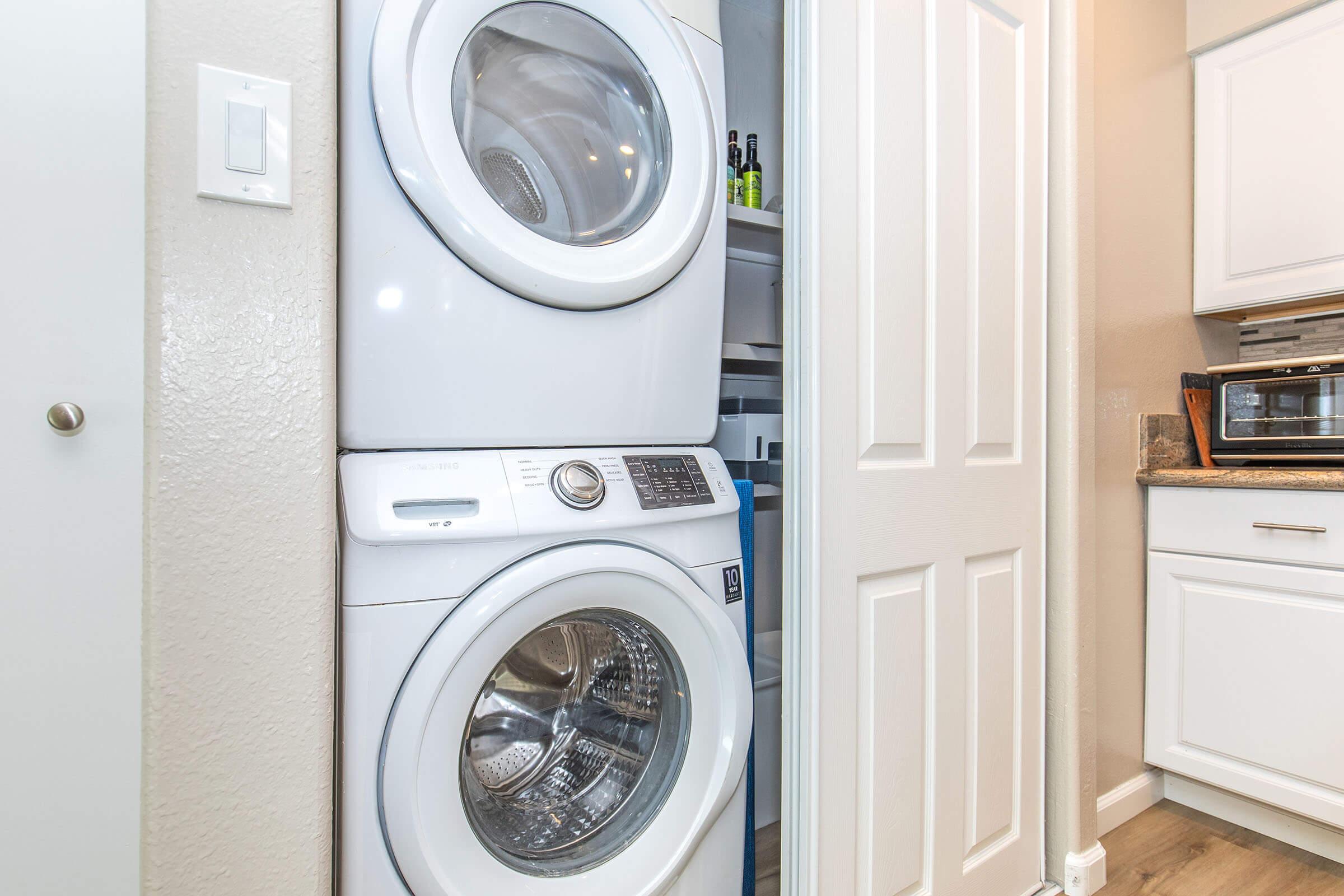 A stackable washer and dryer unit positioned in a narrow laundry space next to a closed pantry door; the surroundings feature light-colored walls, wooden flooring, and modern kitchen cabinetry.