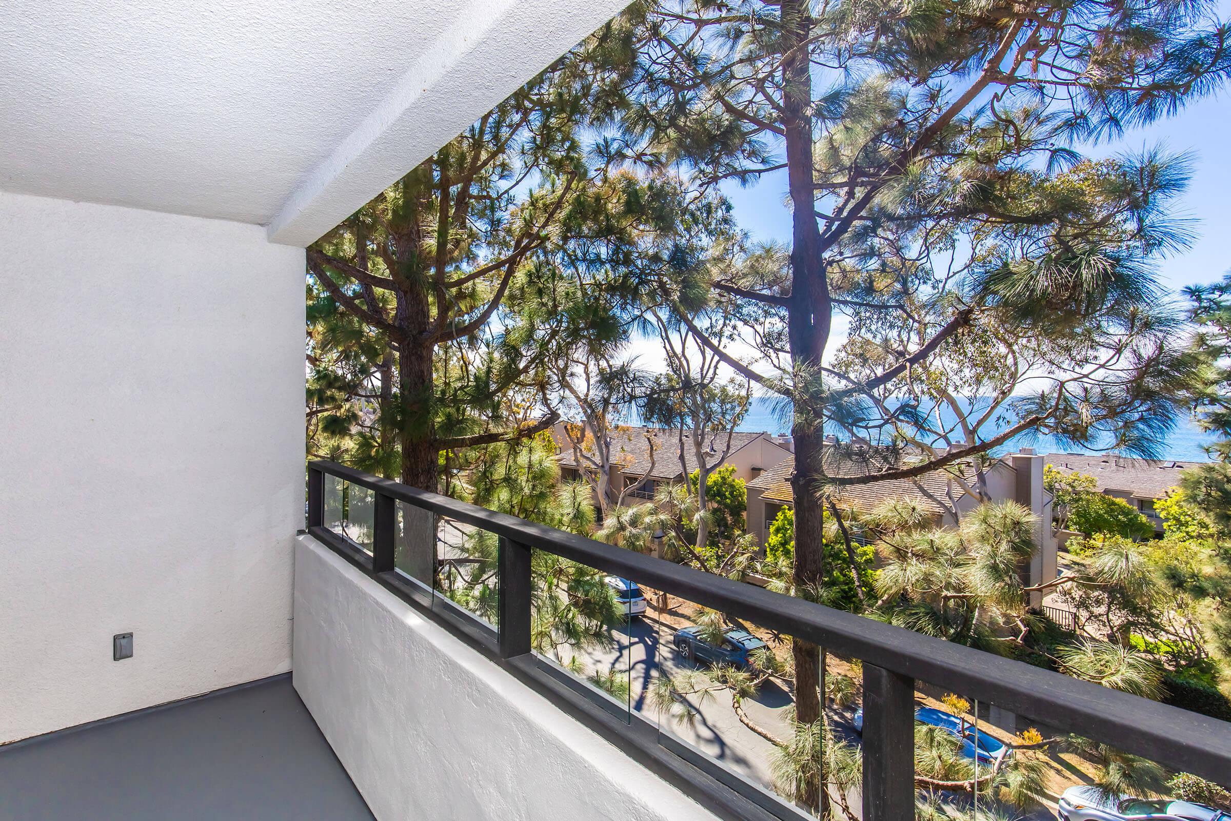 A view from a balcony overlooking trees and rooftops, with a clear blue sky in the background. The balcony features a black railing and white walls, creating a bright and open atmosphere. Sunlight filters through the trees, enhancing the serene outdoor setting.