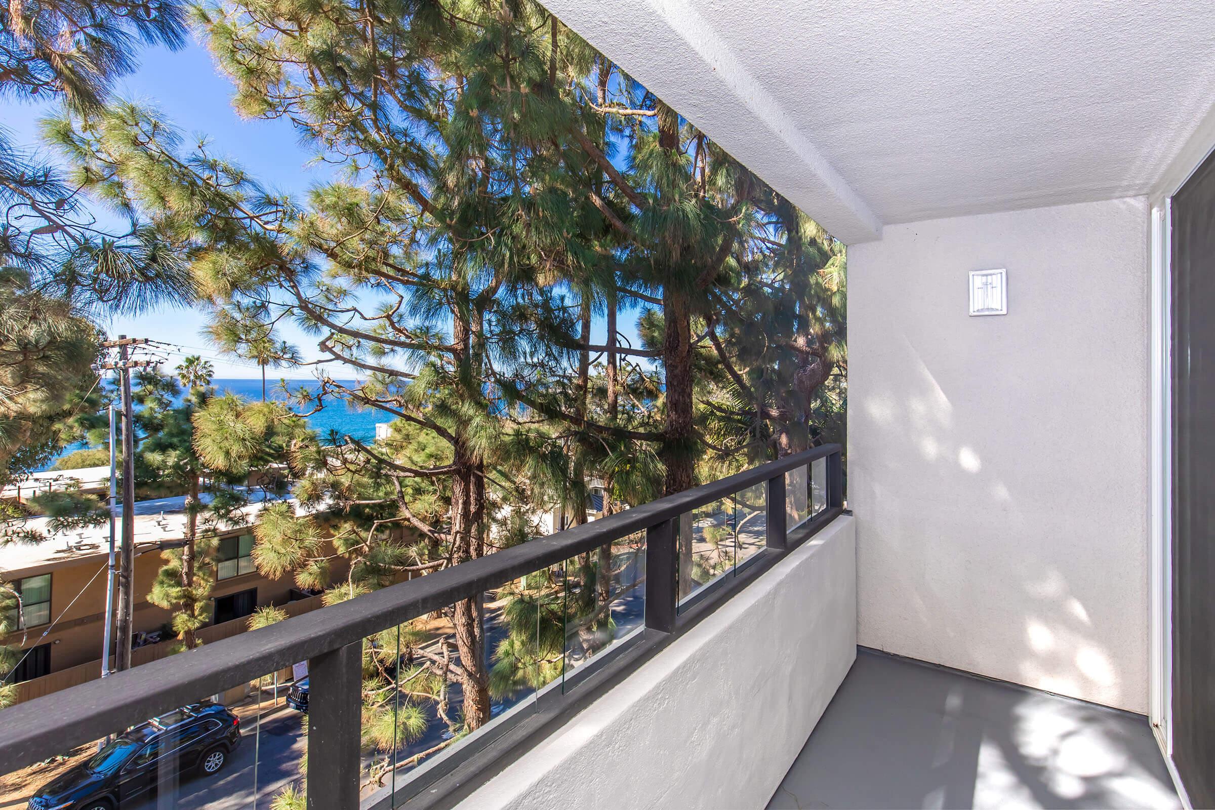 A balcony view featuring tall pine trees and a glimpse of the ocean in the distance. The scene is bright and sunny, showcasing a clear sky and the vibrant colors of nature, along with a modern railing. Ideal for enjoying fresh air and a scenic landscape.