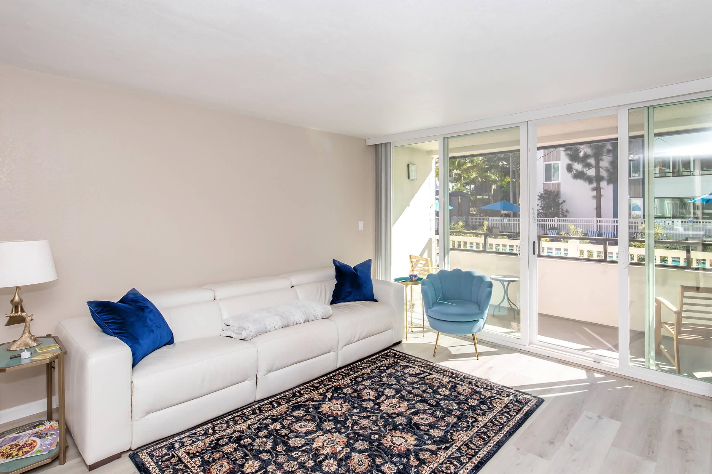 A modern living room featuring a white sofa with blue pillows, a small side table with a lamp, and a decorative patterned rug. Large glass sliding doors lead to a balcony or patio area with outdoor seating visible. The walls are painted a light color, creating a bright and open atmosphere.