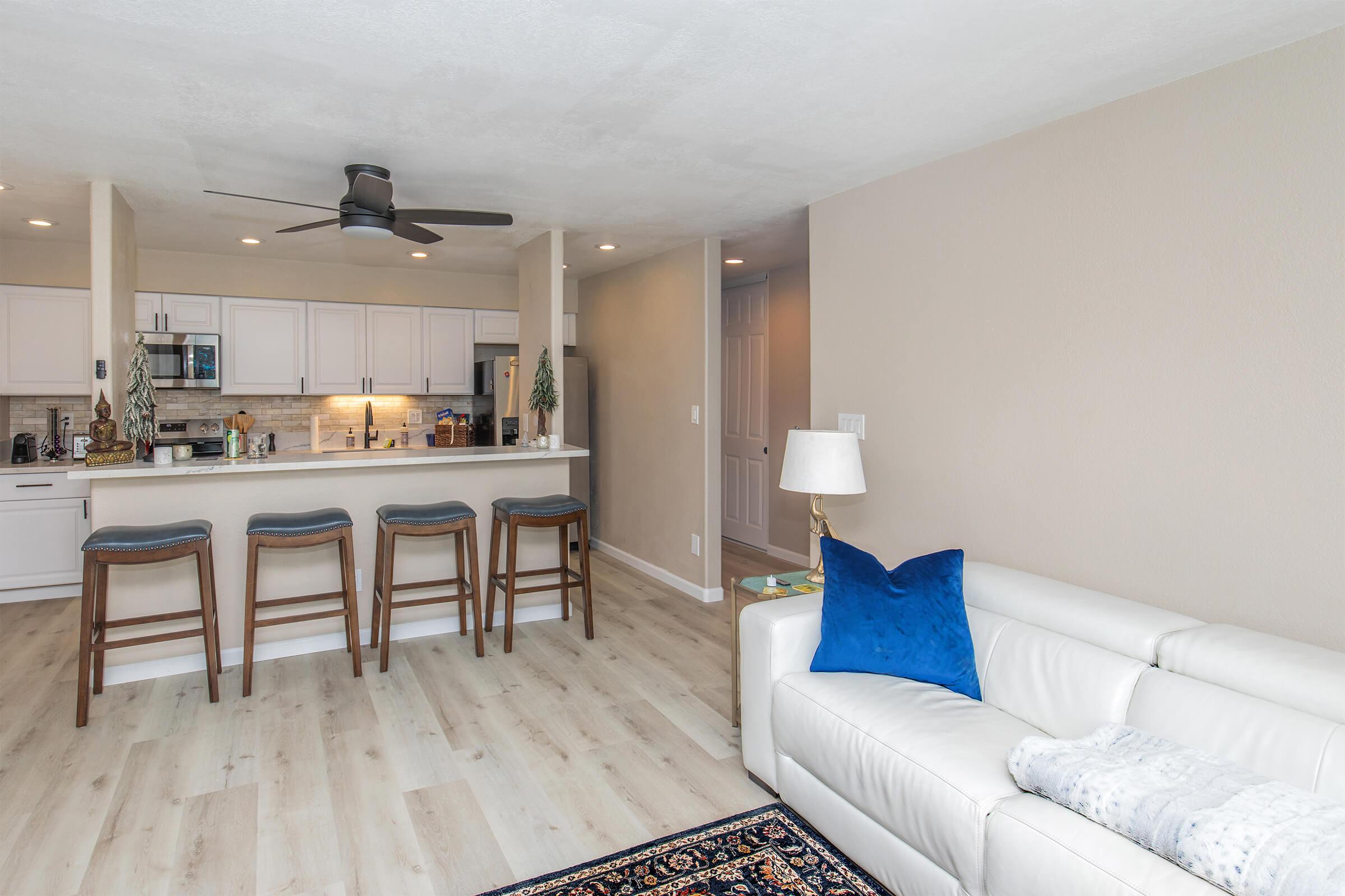 Bright and modern living space featuring a light-colored sofa with a blue accent pillow, a wooden dining area with four high stools, and a kitchen with white cabinets and stainless steel appliances. Light wood flooring and neutral wall colors create a warm, inviting atmosphere.