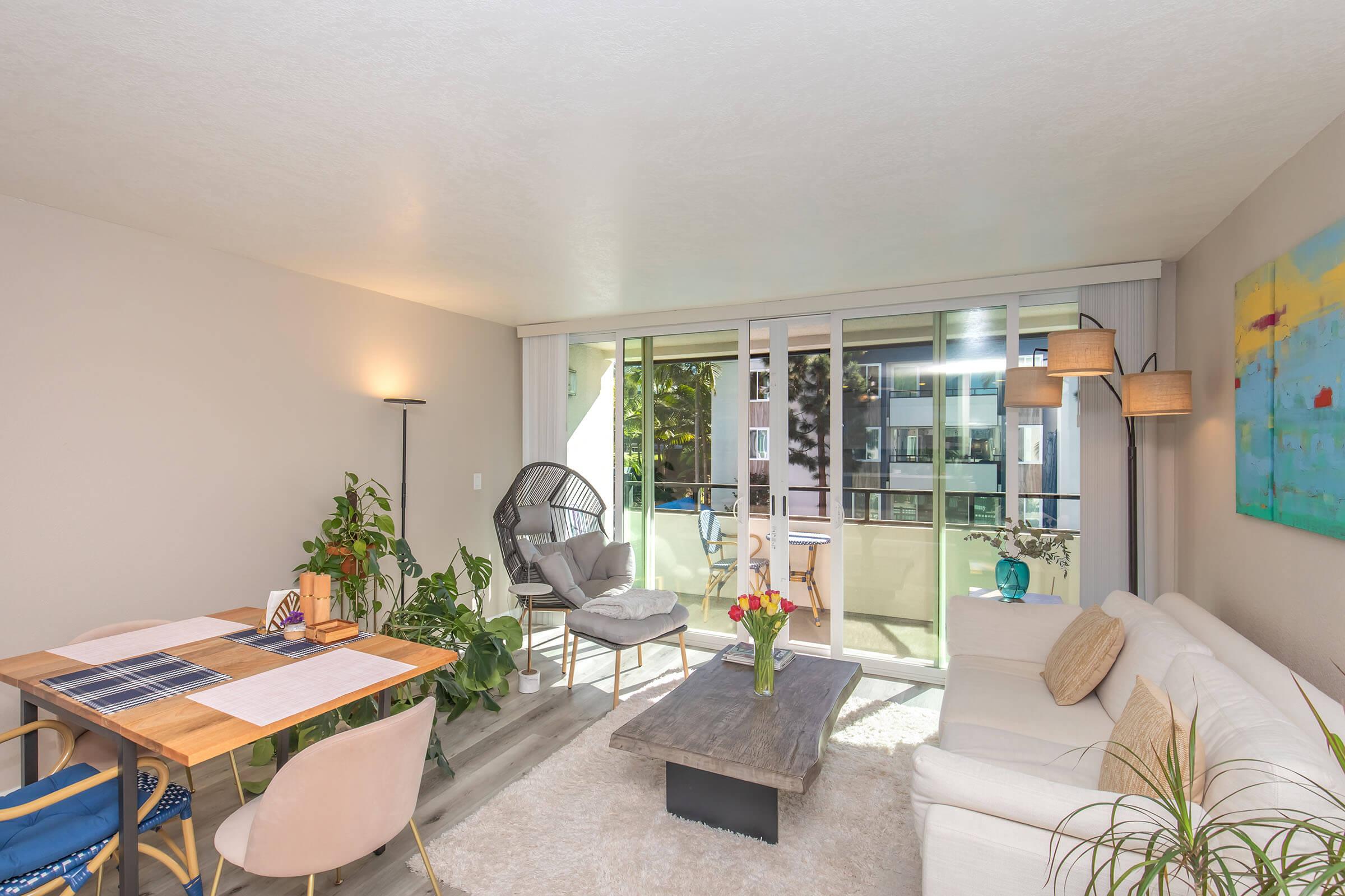 A bright and modern living room featuring a white sofa, a wooden coffee table, and a dining area with a table and chairs. Large windows provide natural light and a view of a balcony with greenery. Decor includes potted plants and abstract art on the walls.