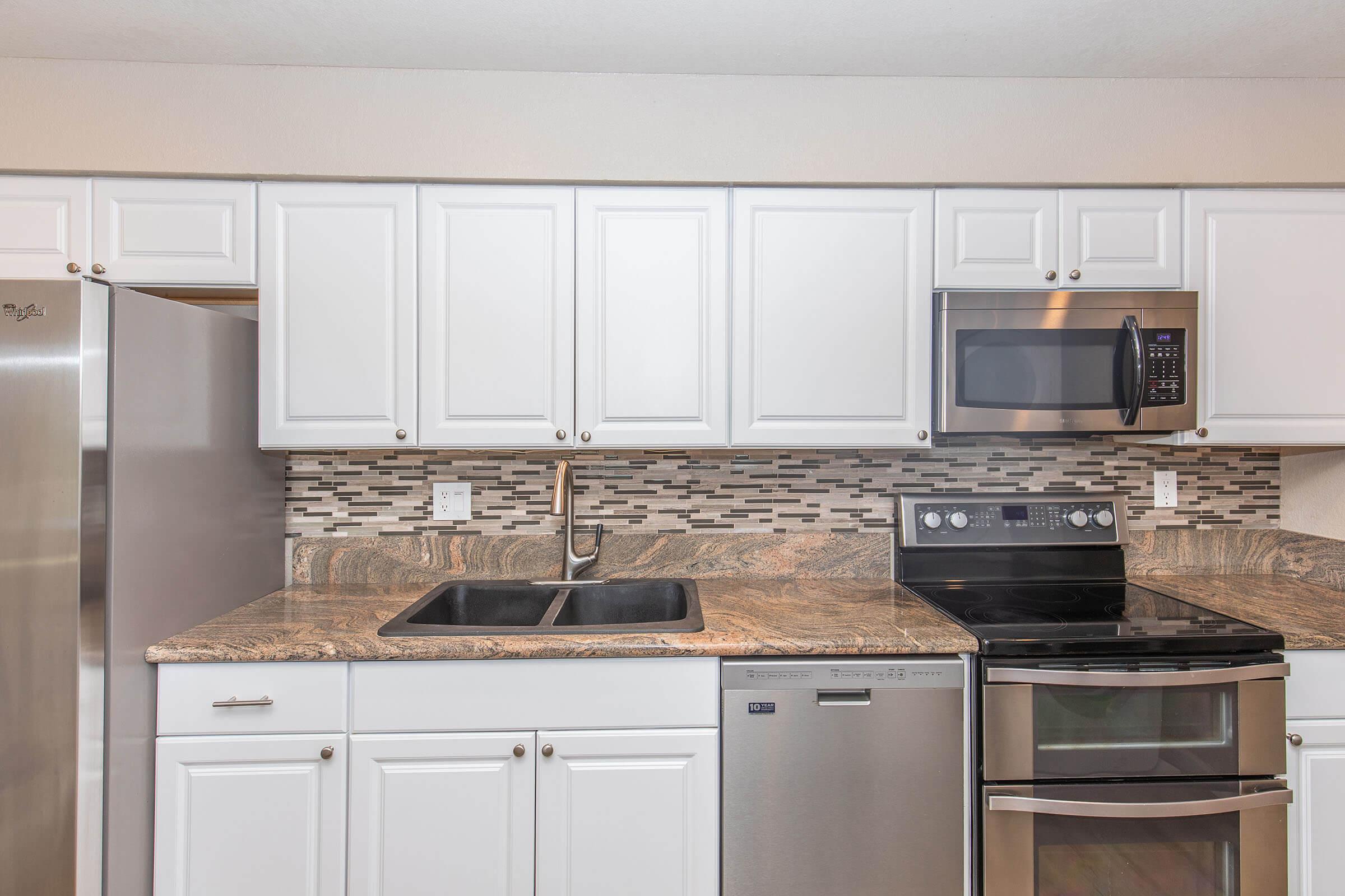 Modern kitchen featuring white cabinetry, a dual sink, stainless steel appliances, including a microwave and an electric oven with a glass cooktop. The countertop has a natural stone appearance, and a decorative tile backsplash adds detail, enhancing the overall contemporary design.