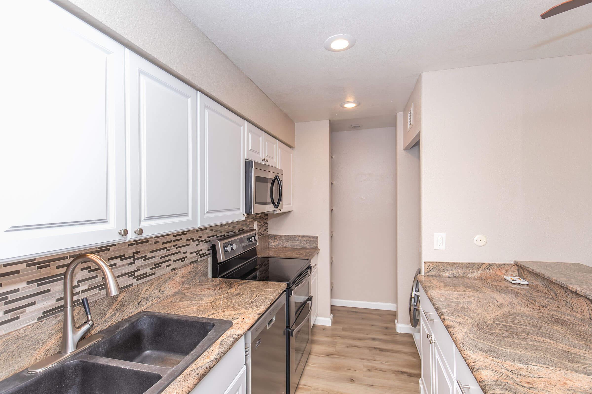 Modern kitchen featuring white cabinetry, dark countertops, stainless steel appliances, including a microwave and oven, and a double sink. The backsplash is made of varied tiles. The flooring is light wood, and the space is well-lit with recessed lighting.