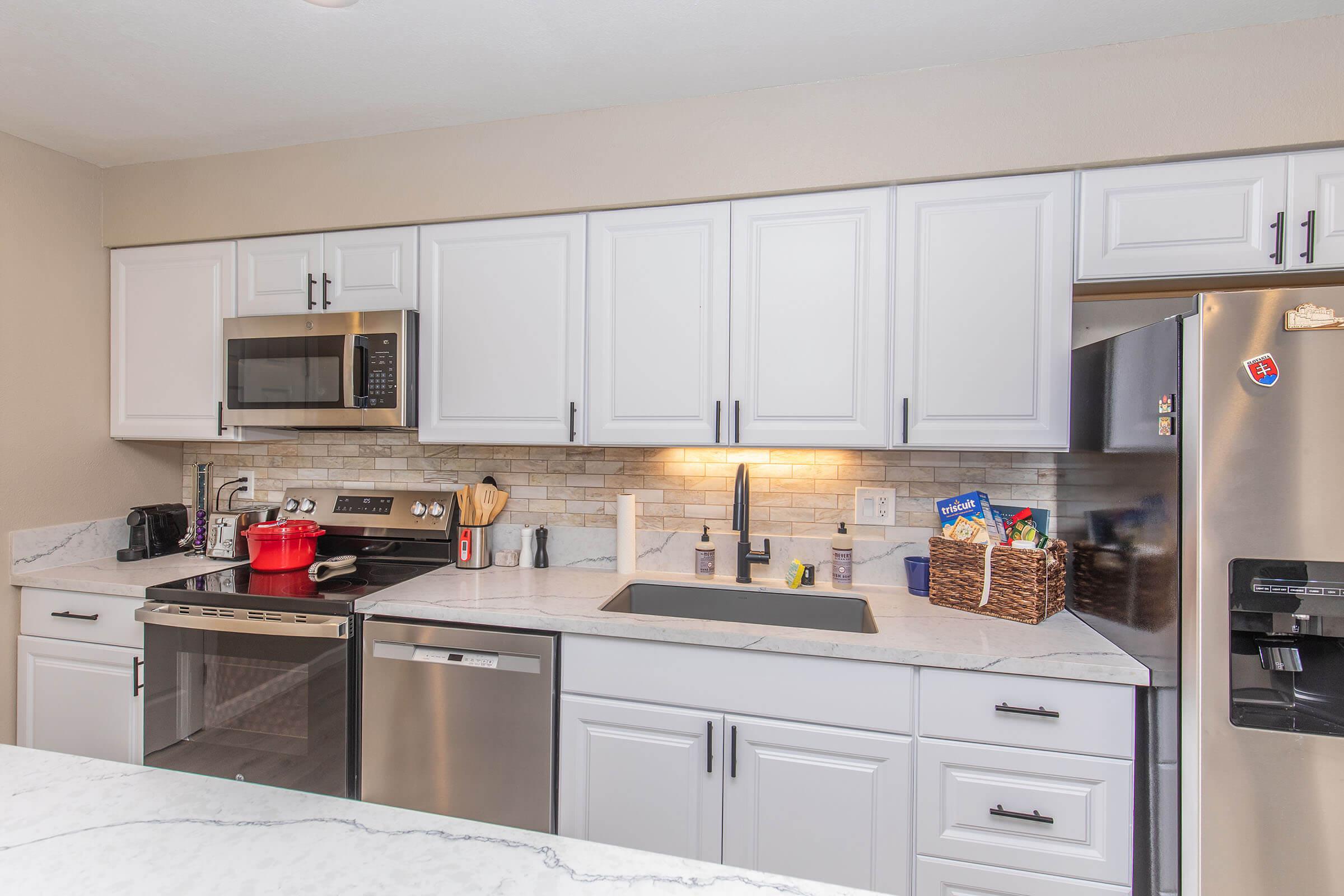 A modern kitchen featuring white cabinetry, stainless steel appliances, and a marble countertop. The setup includes a microwave, oven, refrigerator, and a sink. There are kitchen utensils, a red pot, and a wicker basket on the counter, creating a functional and stylish cooking space.