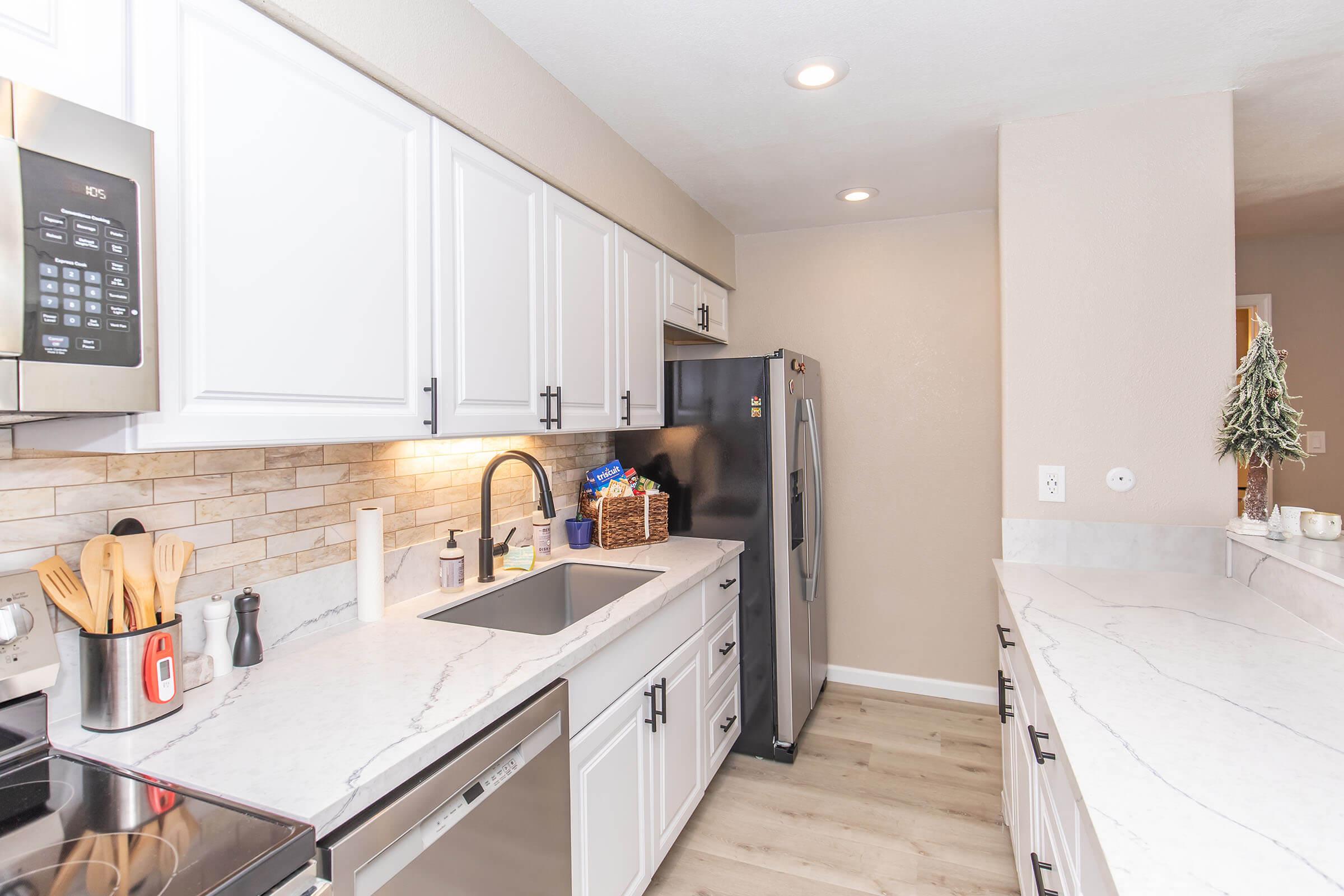 A modern kitchen featuring white cabinets, a stainless steel refrigerator, a silver dishwasher, and a marble countertop. The space includes a sink with a decorative backsplash, wooden cooking utensils, and soft lighting, creating a bright and inviting atmosphere.