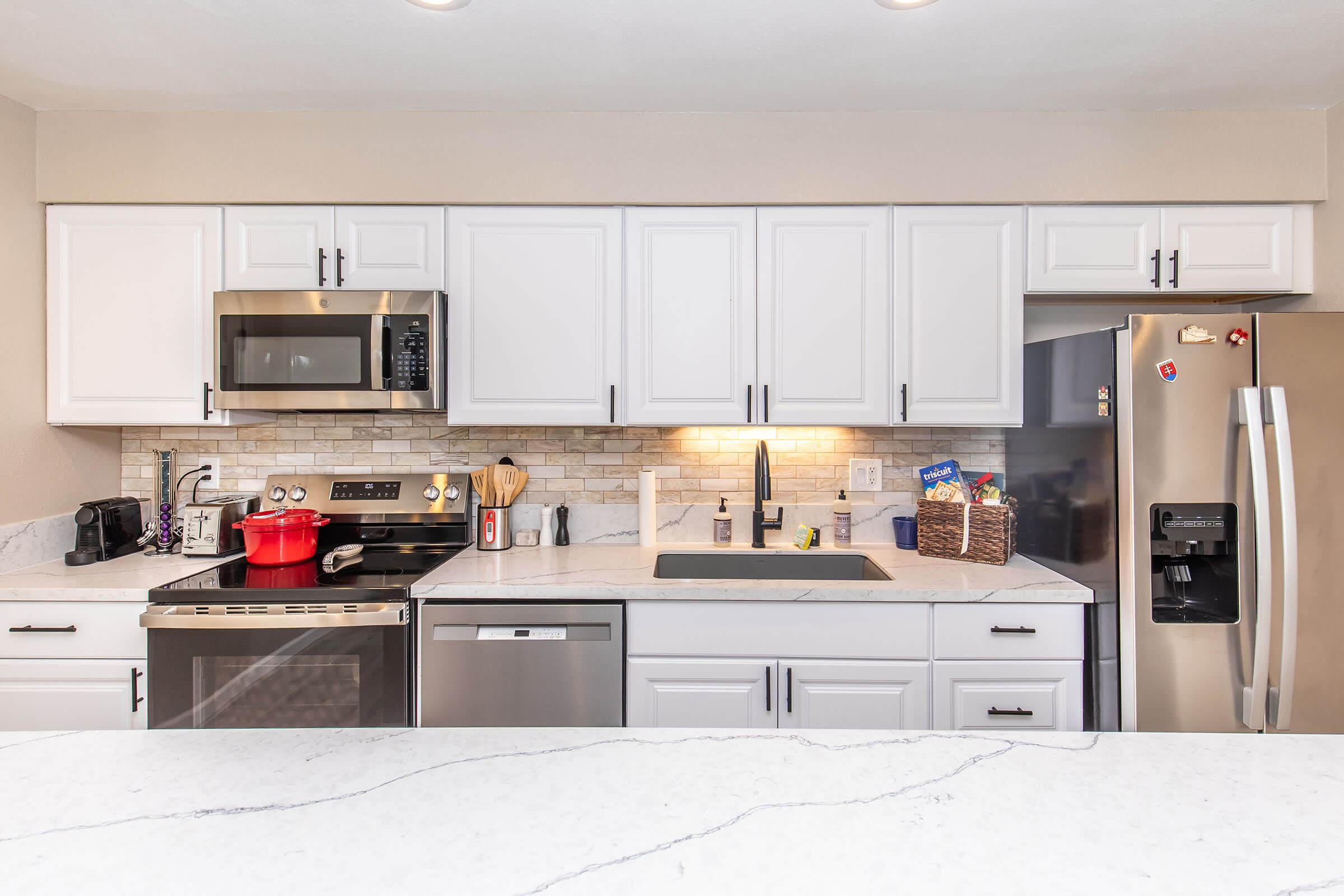 A modern kitchen featuring white cabinetry, a stainless steel refrigerator, and an oven with a microwave above. The countertop is white with gray veining, and a red pot sits on the stove. There are various kitchen utensils and a sink integrated into the countertop. Soft lighting highlights the space.