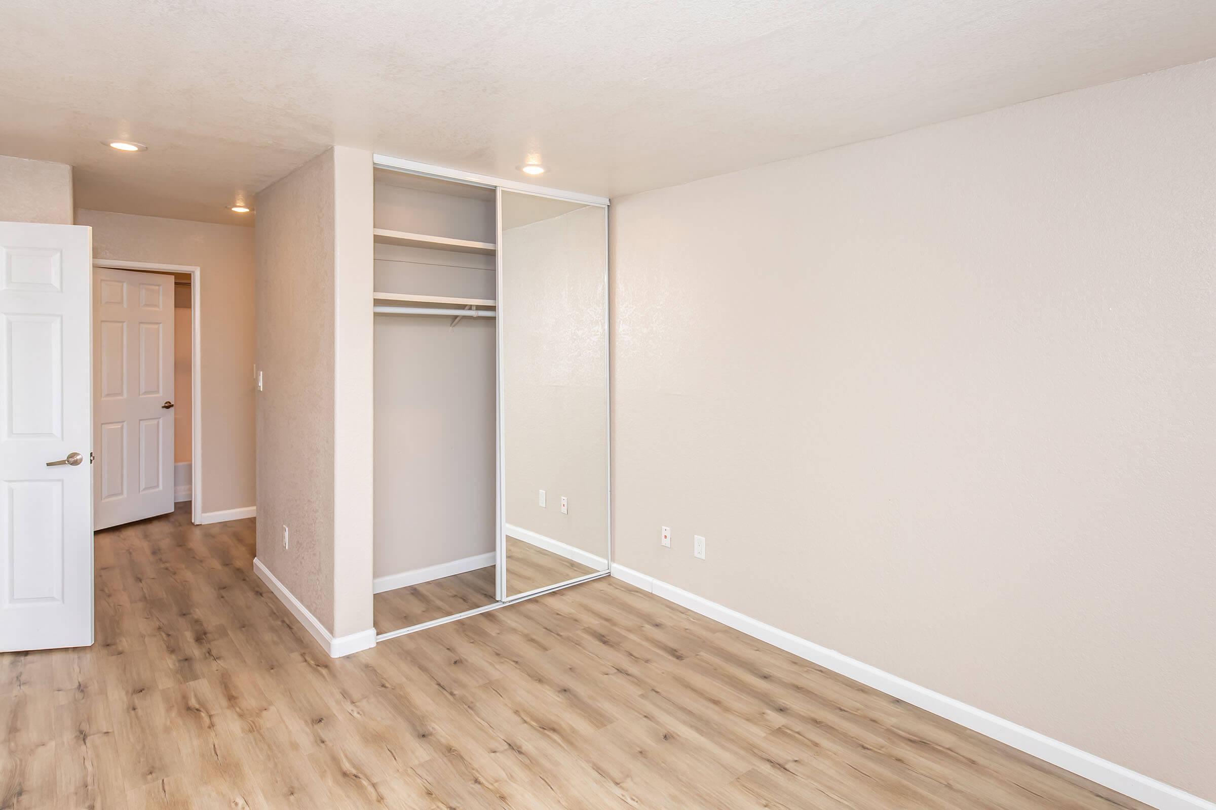 Empty room with light-colored walls and a mirrored closet. The flooring is a light wood finish. There is a door on the left leading to another room. The space is well-lit with ceiling lights, creating a bright and airy atmosphere.