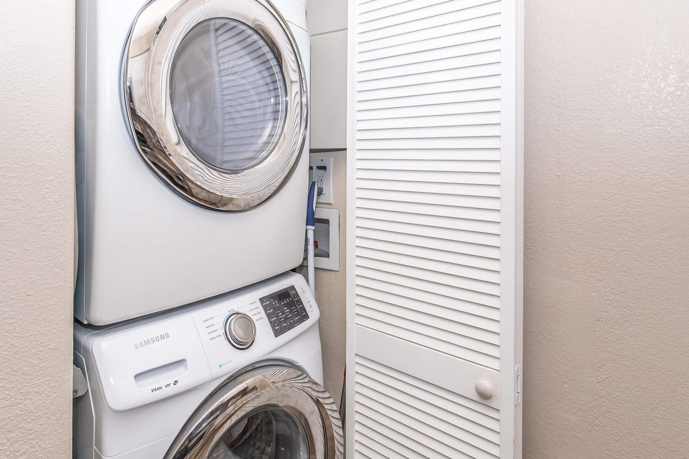 A stackable washer and dryer unit in a small laundry space, with a light-colored wall and a closed louvered door nearby. The appliances have a modern design with chrome accents.
