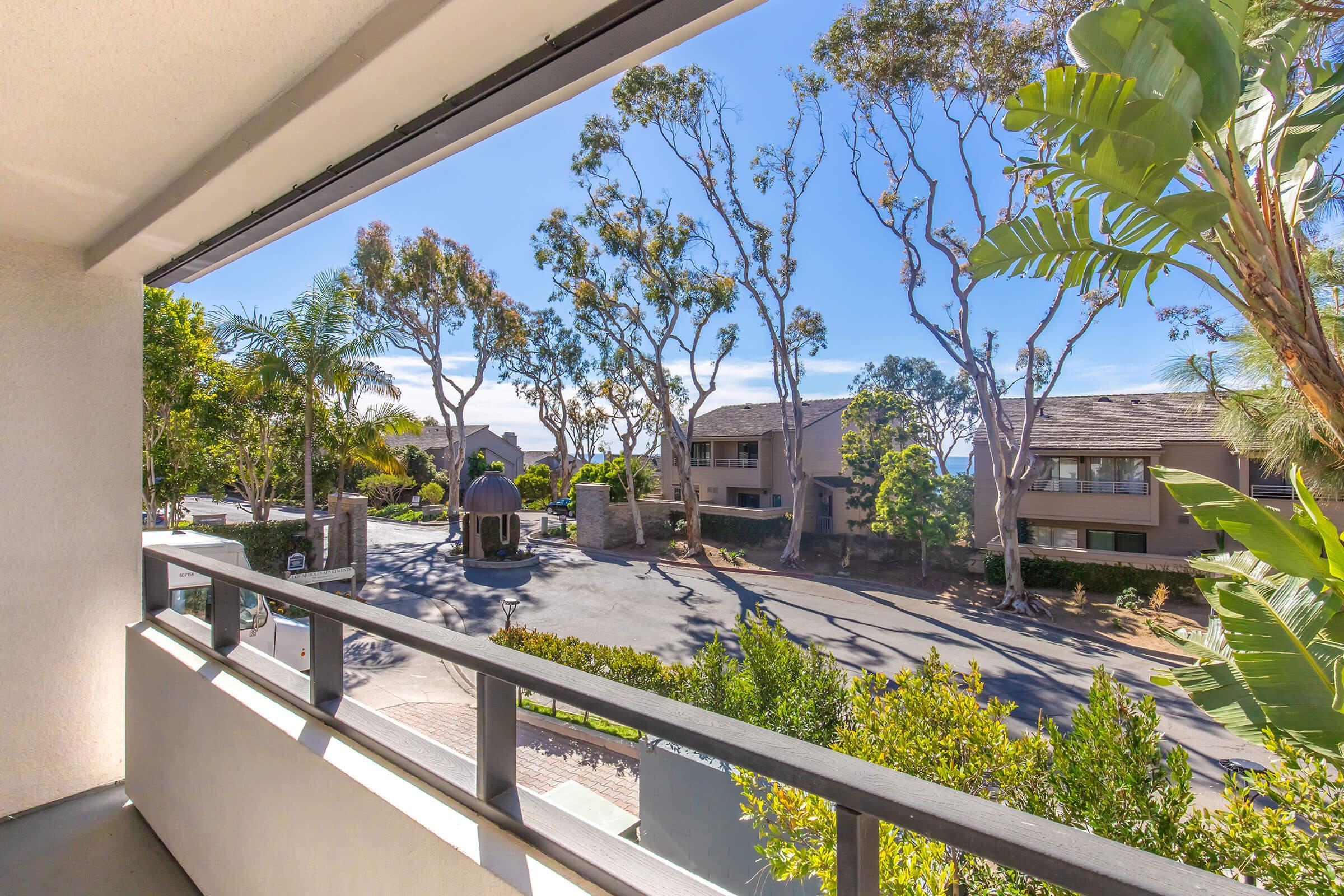 View from a balcony overlooking a landscaped area with trees and shrubs. The scene includes residential buildings in the background and a pathway leading towards a gazebo or outdoor structure. The sky is clear, suggesting a sunny day.