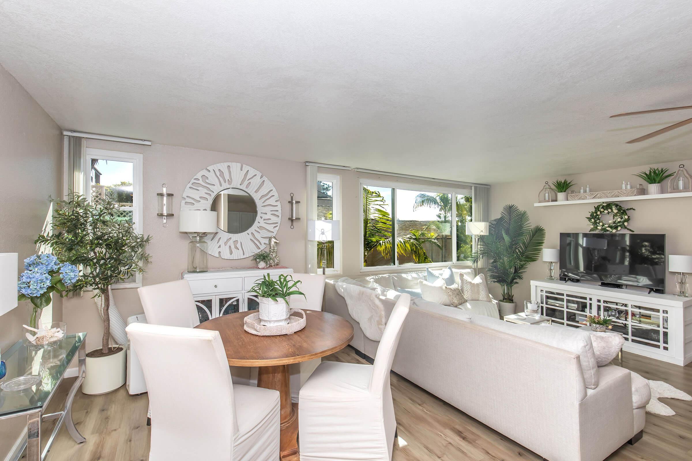 A bright and airy living room featuring a white sofa, glass coffee table, and circular mirror. A round wooden dining table with white chairs is nearby. The space is decorated with potted plants and has large windows allowing natural light, creating a fresh and inviting atmosphere.