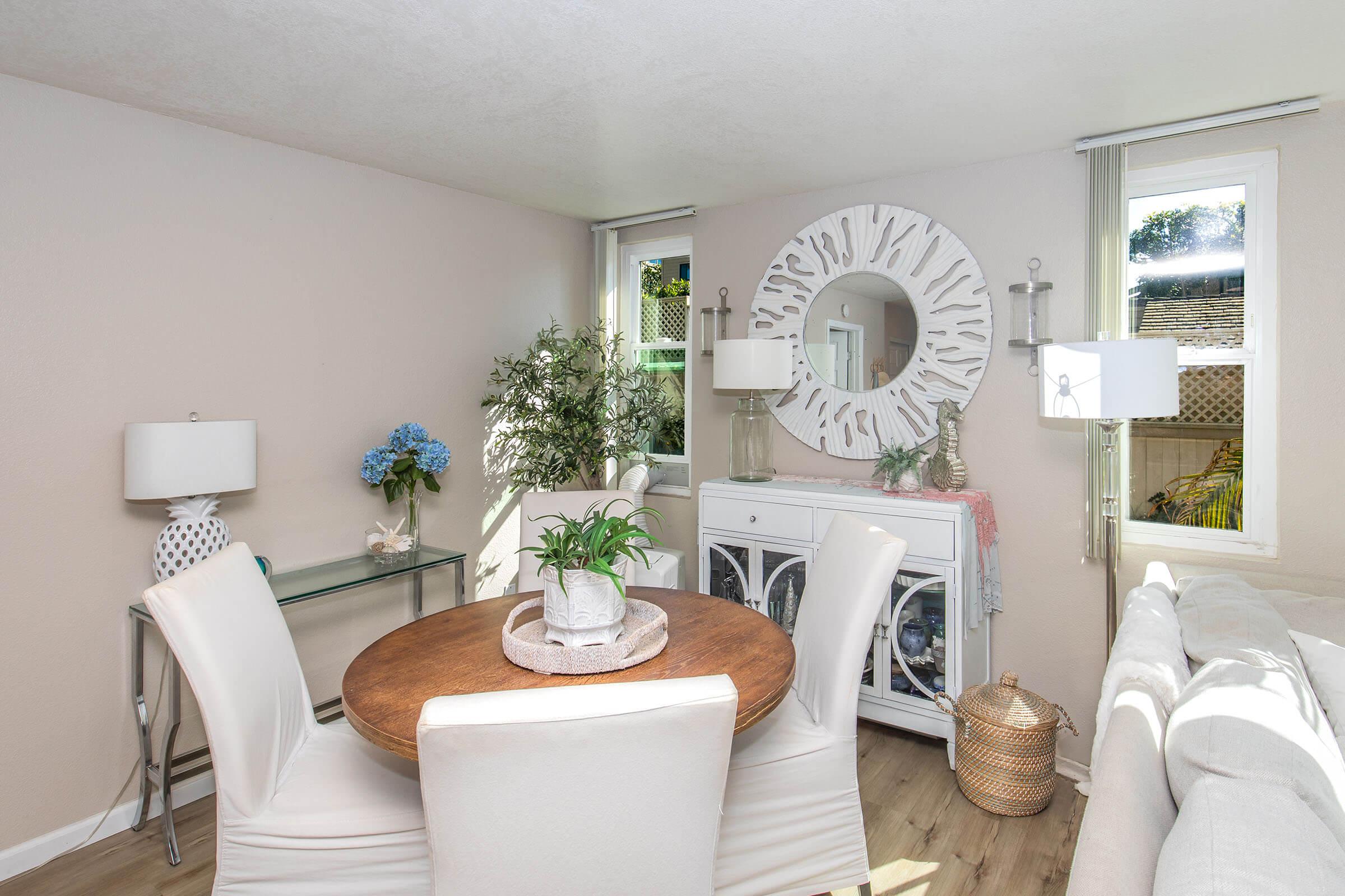 A cozy living space featuring a round wooden dining table with white chairs, a decorative mirror on the wall, and a sideboard. Potted plants and blue flowers add color to the room, complemented by two lamps. Large windows allow natural light to filter in, creating a bright and inviting atmosphere.