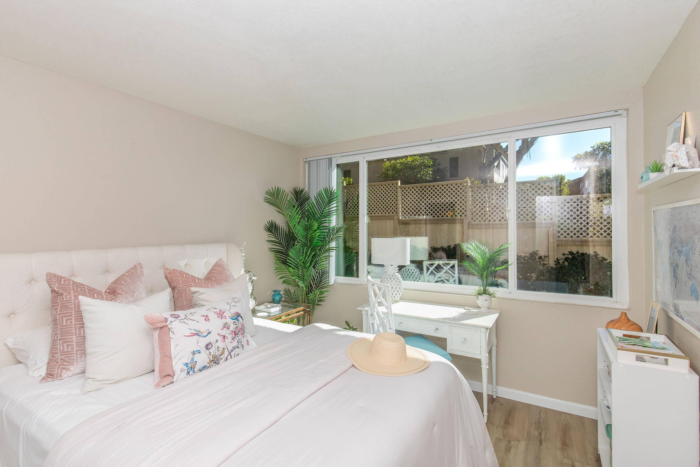 A cozy bedroom featuring a light-colored bed with decorative pillows, a small desk with a chair, and a large window allowing natural light to enter. Green plants are positioned near the window, providing a fresh and inviting atmosphere. Light wood flooring complements the soft color scheme.