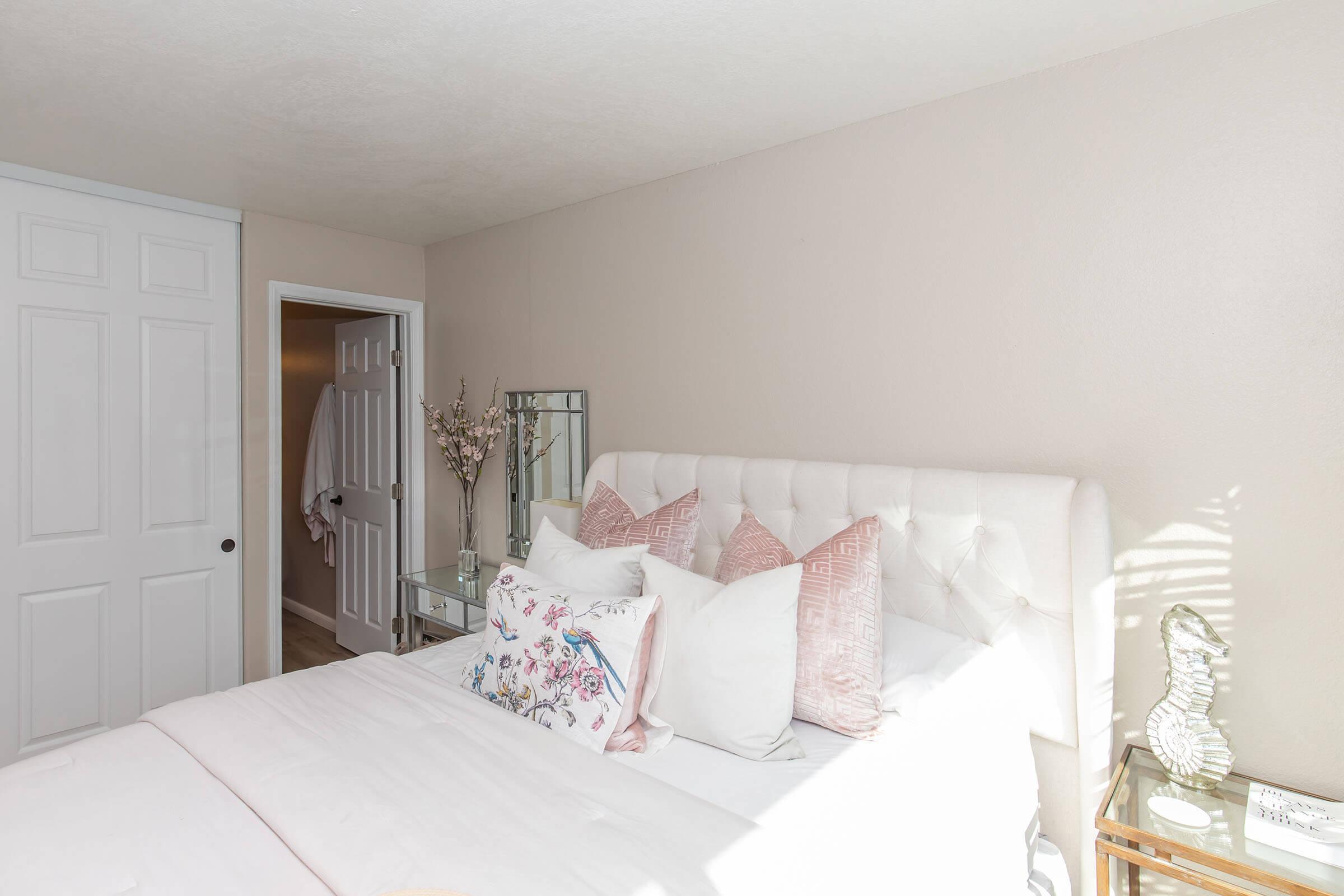 A bright and cozy bedroom featuring a tufted white headboard with decorative pillows. A mirrored nightstand holds a decorative vase, and there's a glimpse of a white door leading to another room. Soft natural light enhances the serene atmosphere, complemented by light-colored walls.