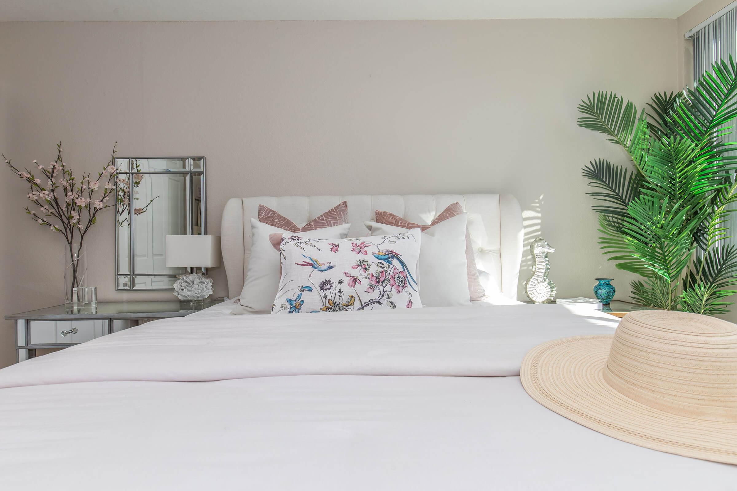 A neatly arranged bedroom featuring a white bed with decorative pillows, a large straw hat resting on the bed, a mirrored nightstand with a lamp, and a small flowering branch. A decorative palm plant adds a touch of greenery to the room, enhancing the serene and inviting atmosphere.