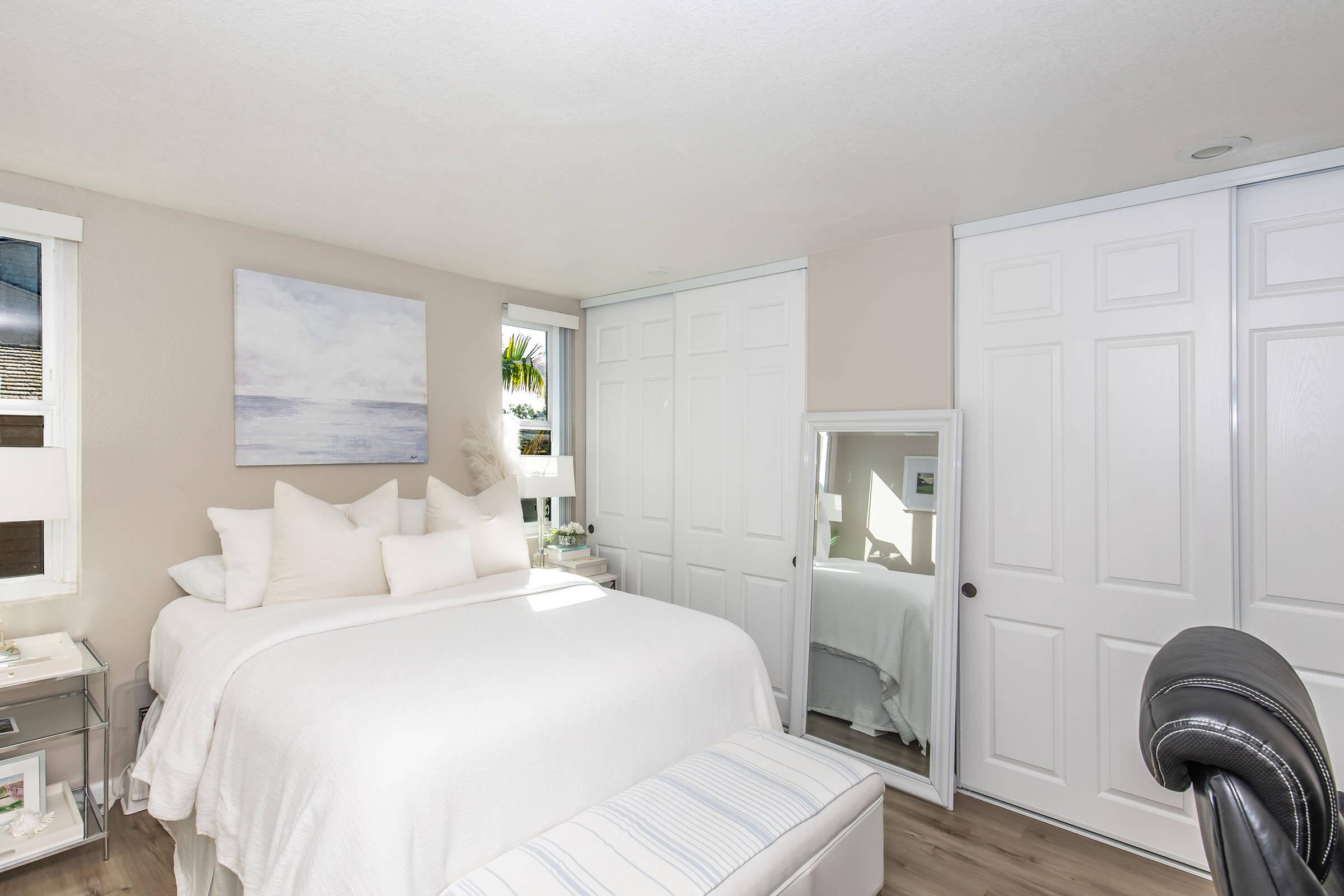 A cozy, well-lit bedroom featuring a white bed with multiple pillows. A large mirror sits beside the bed, and white closet doors are visible in the background. The room has a serene, neutral color palette, complemented by a piece of artwork depicting a calm ocean scene on the wall.