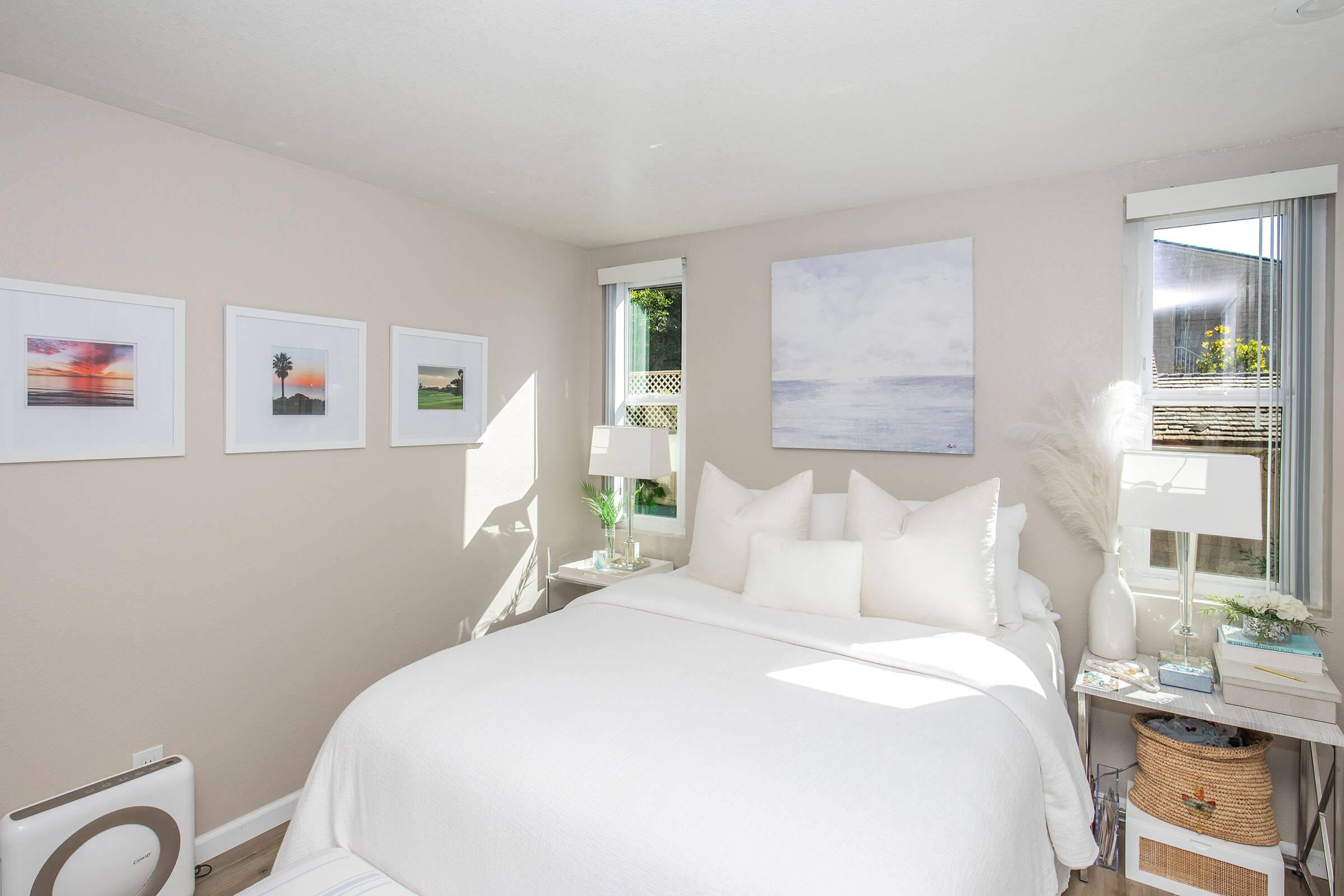 Bright and airy bedroom featuring a white bed with multiple pillows, two stylish bedside lamps, and framed artwork on the walls. Large windows allow natural light to fill the room, enhancing the serene atmosphere. A small table with decorative items adds charm to the cozy space.