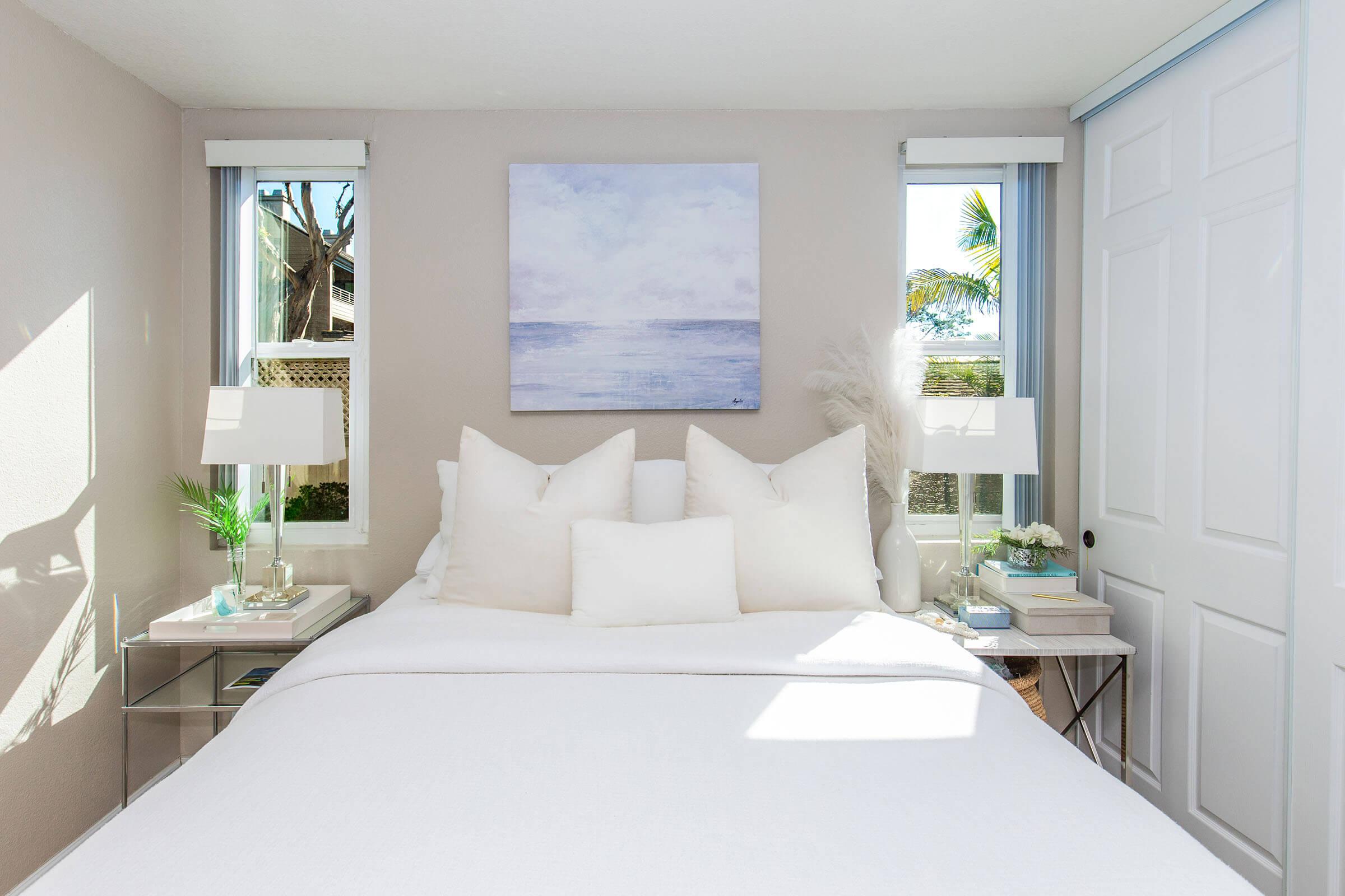 A bright, airy bedroom featuring a white bed with multiple pillows, silver bedside tables with decorative plants and books. A soft ocean-themed artwork hangs above the bed. Natural light streams in from two windows, illuminating the space and highlighting the neutral color palette.