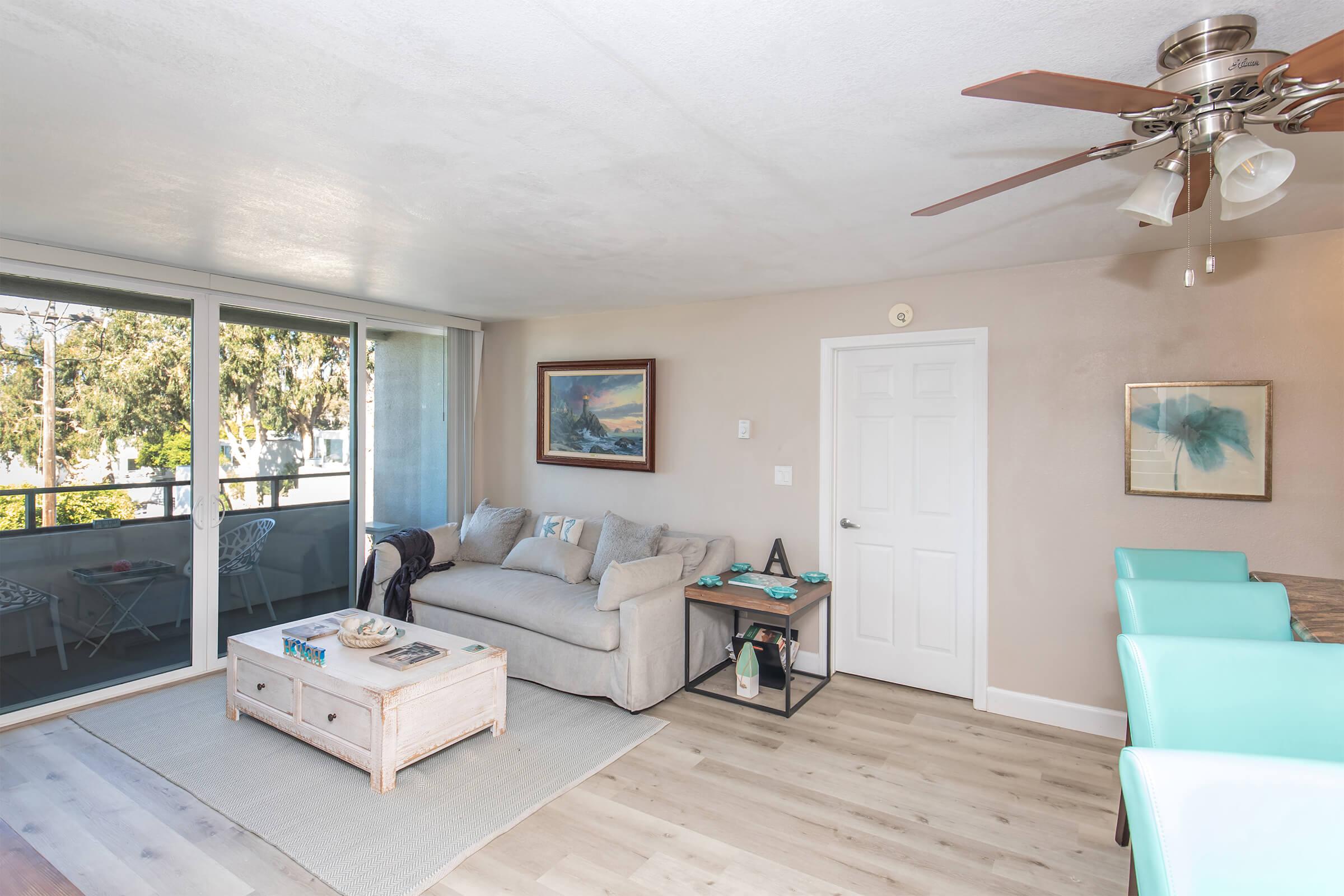 Bright and airy living room featuring a light-colored sofa, a coffee table, and a ceiling fan. A glass door leads to a balcony with outdoor seating. Light wood flooring and soft wall colors create a welcoming atmosphere. Decor includes artwork and a small side table.