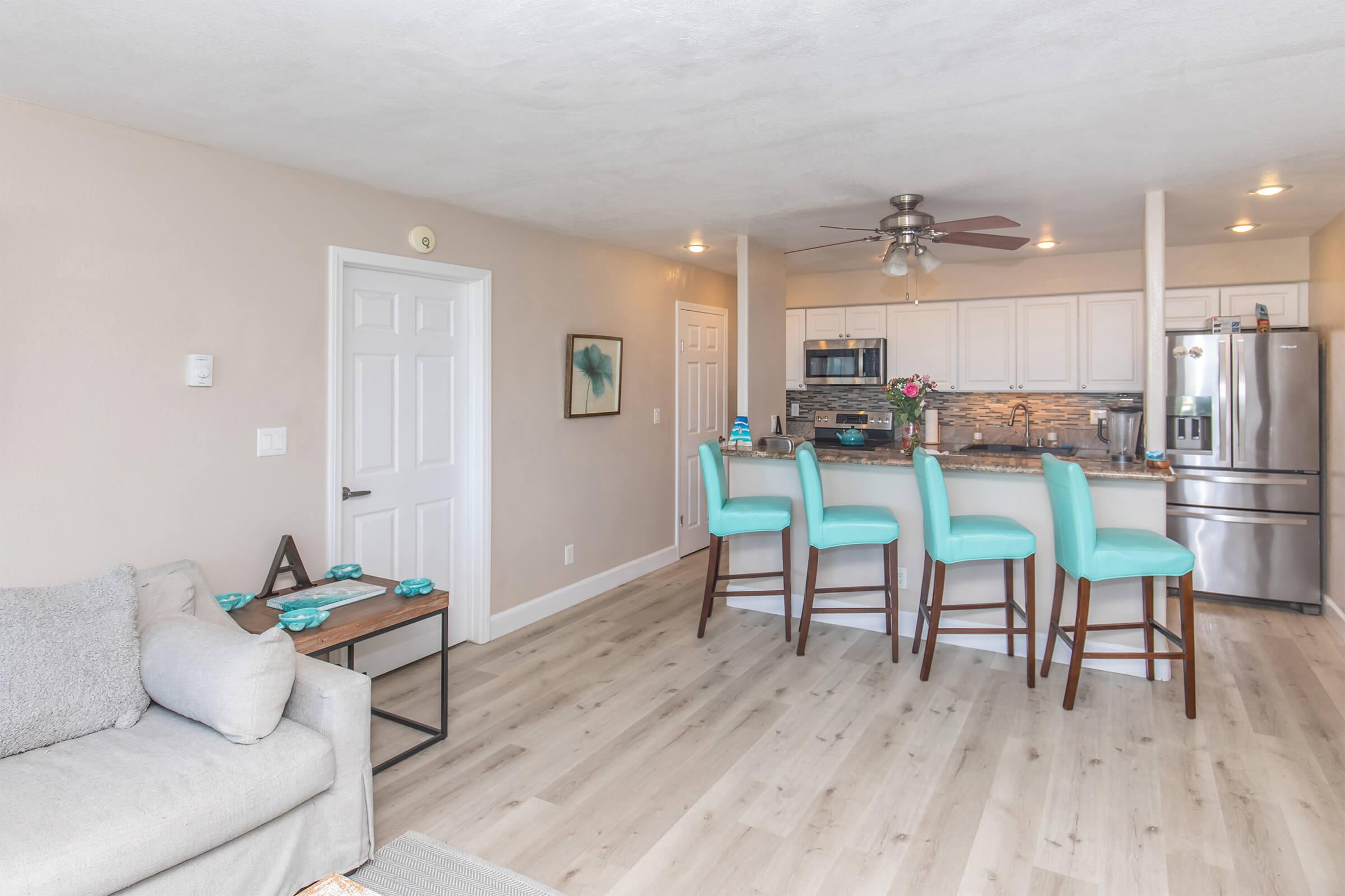 Bright and modern kitchen and living area featuring light wood flooring, a comfortable gray sofa, and a bar with four turquoise stools. The kitchen has stainless steel appliances, white cabinets, and decorative elements. A door leads to another room, and there is natural light from nearby windows.