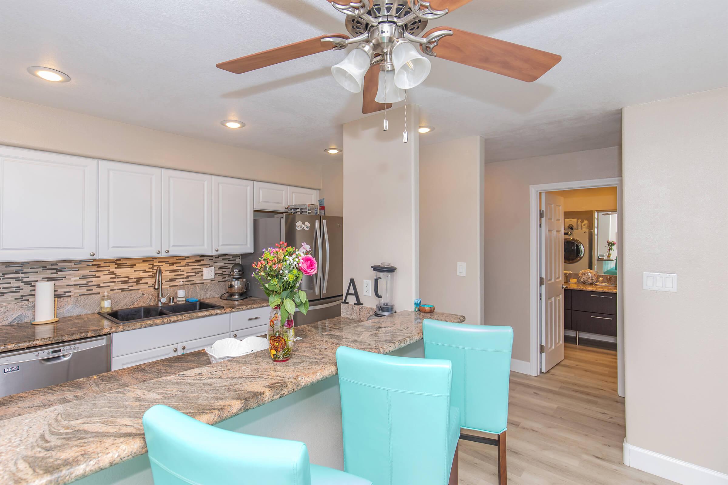 A modern kitchen featuring a granite countertop with bar seating, turquoise stools, and stainless steel appliances. Natural light illuminates the space, which has white cabinetry and a decorative backsplash. A hallway leads to another room, visible on the right. Fresh flowers add a touch of color to the countertop.