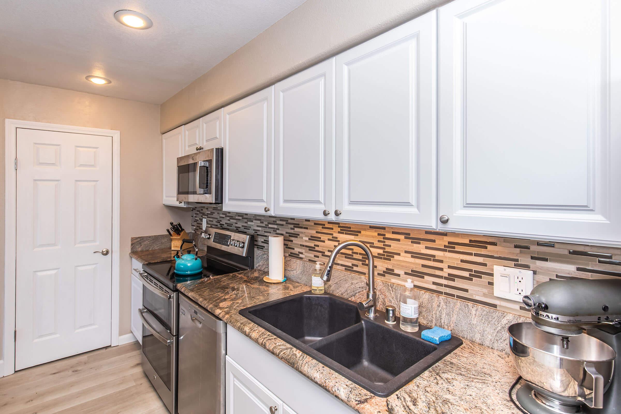 Modern kitchen featuring white cabinets, stainless steel appliances, and a granite countertop. The sink area has a dual-basin design with a faucet and soap dispenser, while the backsplash consists of decorative tiles. A stand mixer is positioned on the countertop next to the sink.