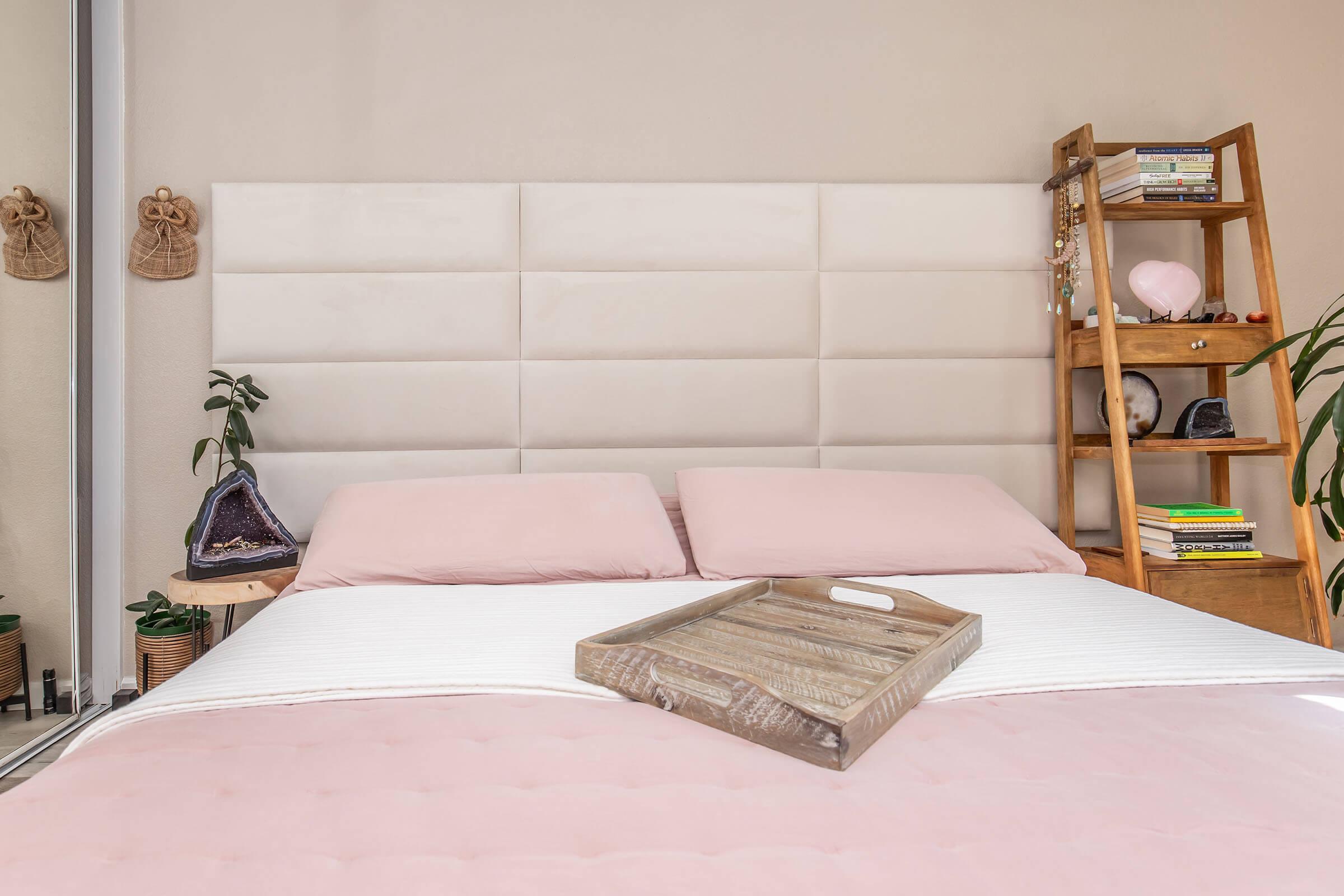 A cozy bedroom featuring a light-colored upholstered headboard above a neatly made bed with pink bedding. A wooden tray sits on the bed. In the background, there's a bookshelf with decorative items and houseplants, creating a warm and inviting atmosphere.