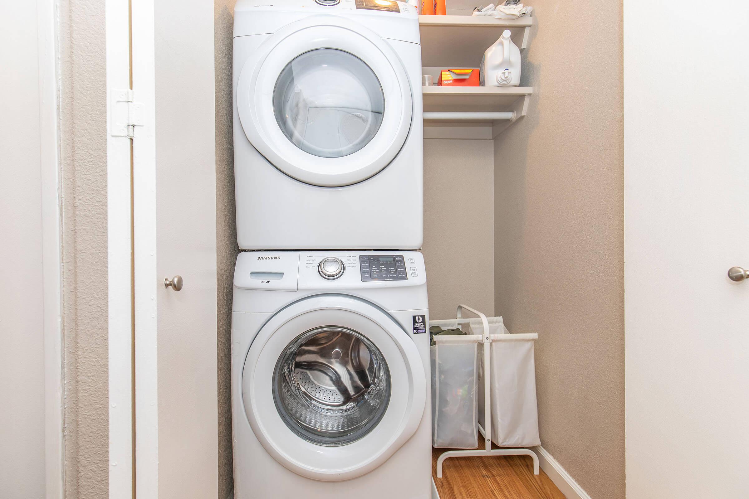 A stacked washer and dryer located in a small laundry closet. The appliances are white, with the dryer on top and the washing machine below. A shelf above the machines holds various laundry supplies, while a laundry basket is situated nearby. The walls are painted in a neutral tone.