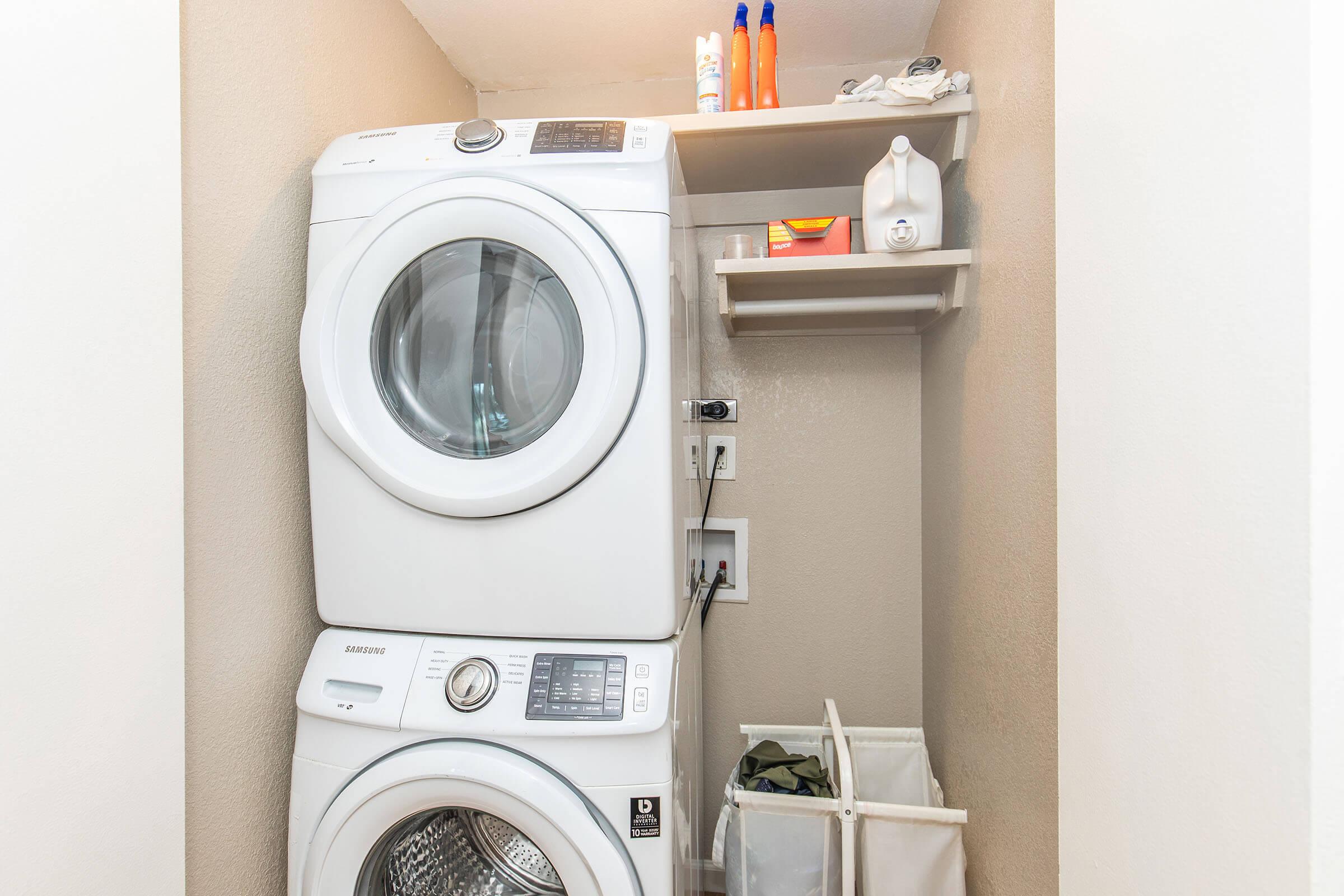 A compact laundry area featuring a stacked white washer and dryer, with a shelf above holding laundry supplies like detergent and fabric softener. To the side, there's a laundry basket with clothes, all set against a neutral-colored wall.