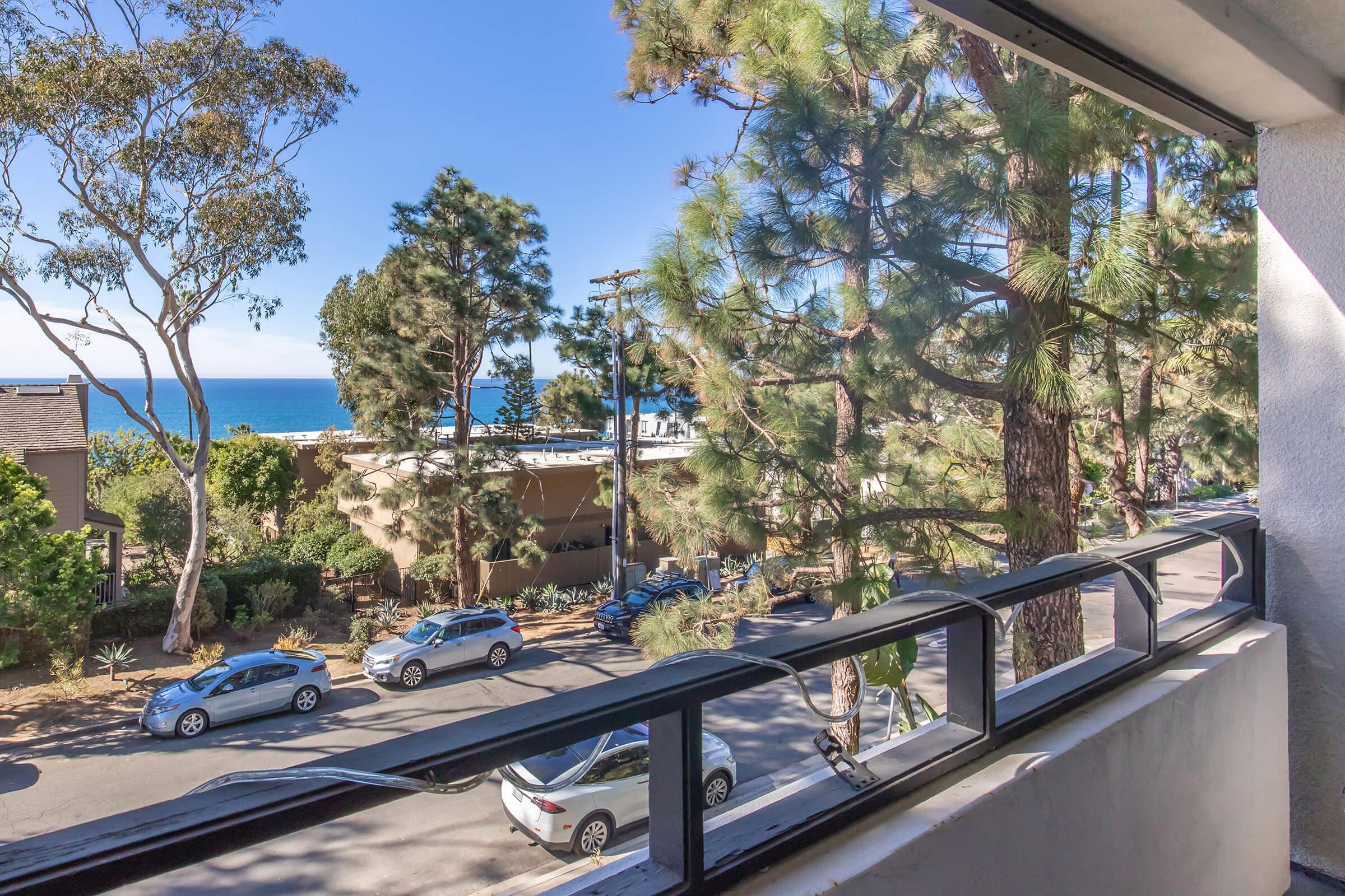 View from a balcony overlooking a street lined with trees, featuring parked cars and a glimpse of the ocean in the distance under a clear blue sky. The scene conveys a serene coastal atmosphere.