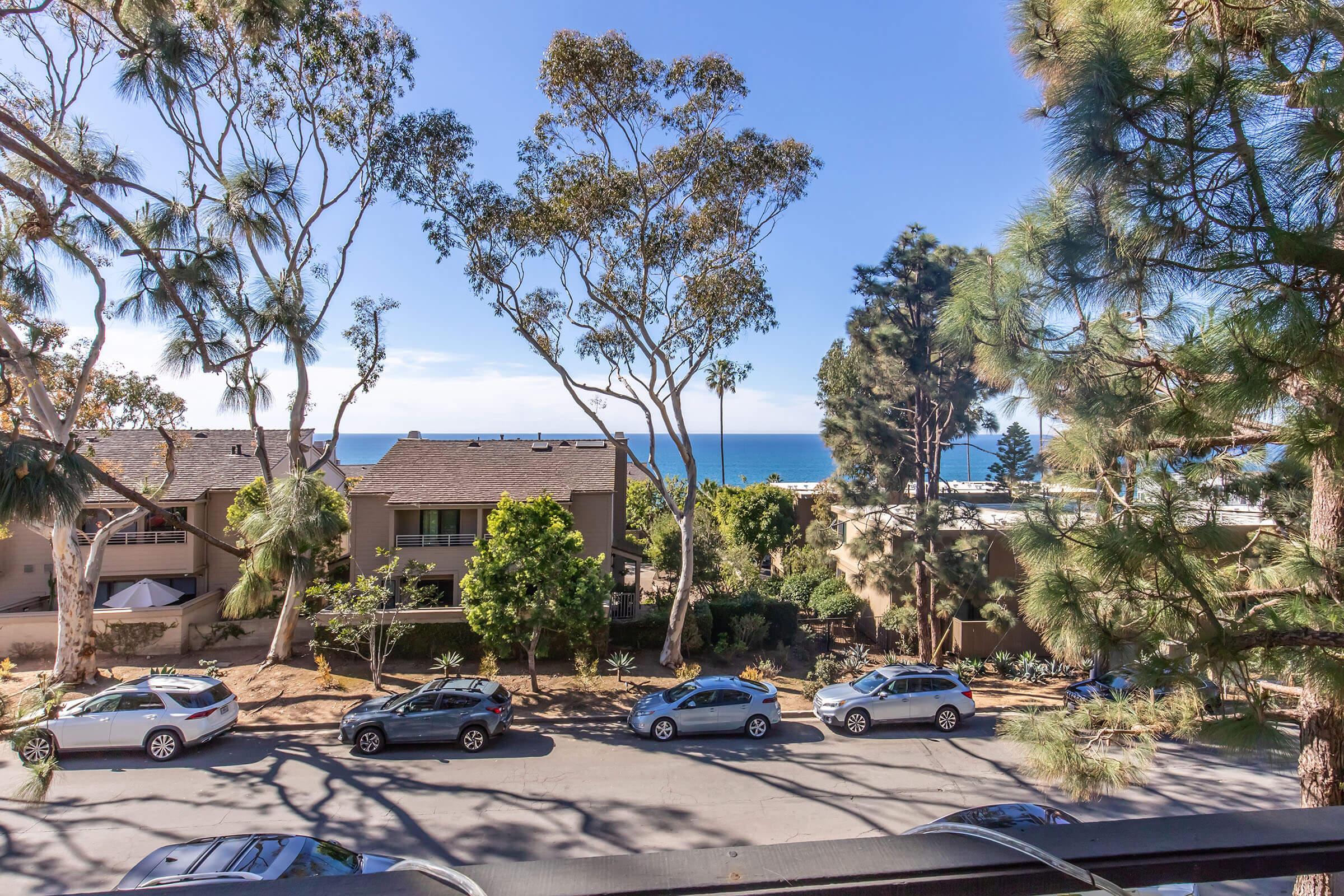 View from a balcony or window showing a coastal scene with palm trees and tall greenery. In the foreground, several cars are parked along a street. In the background, the shimmering blue ocean meets a clear sky, creating a serene and picturesque seaside atmosphere.
