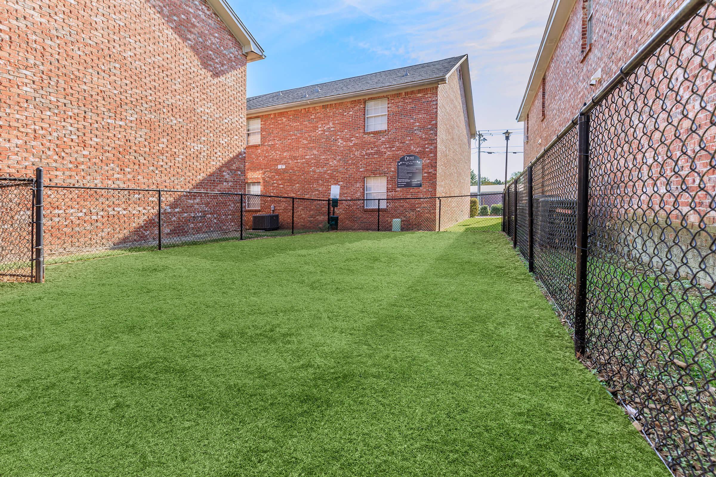 View of a fenced outdoor area between two brick buildings, featuring artificial grass. The space is well-maintained with no obstacles, providing a clean and open environment for activities. In the background, there's a glimpse of utility poles against a blue sky.