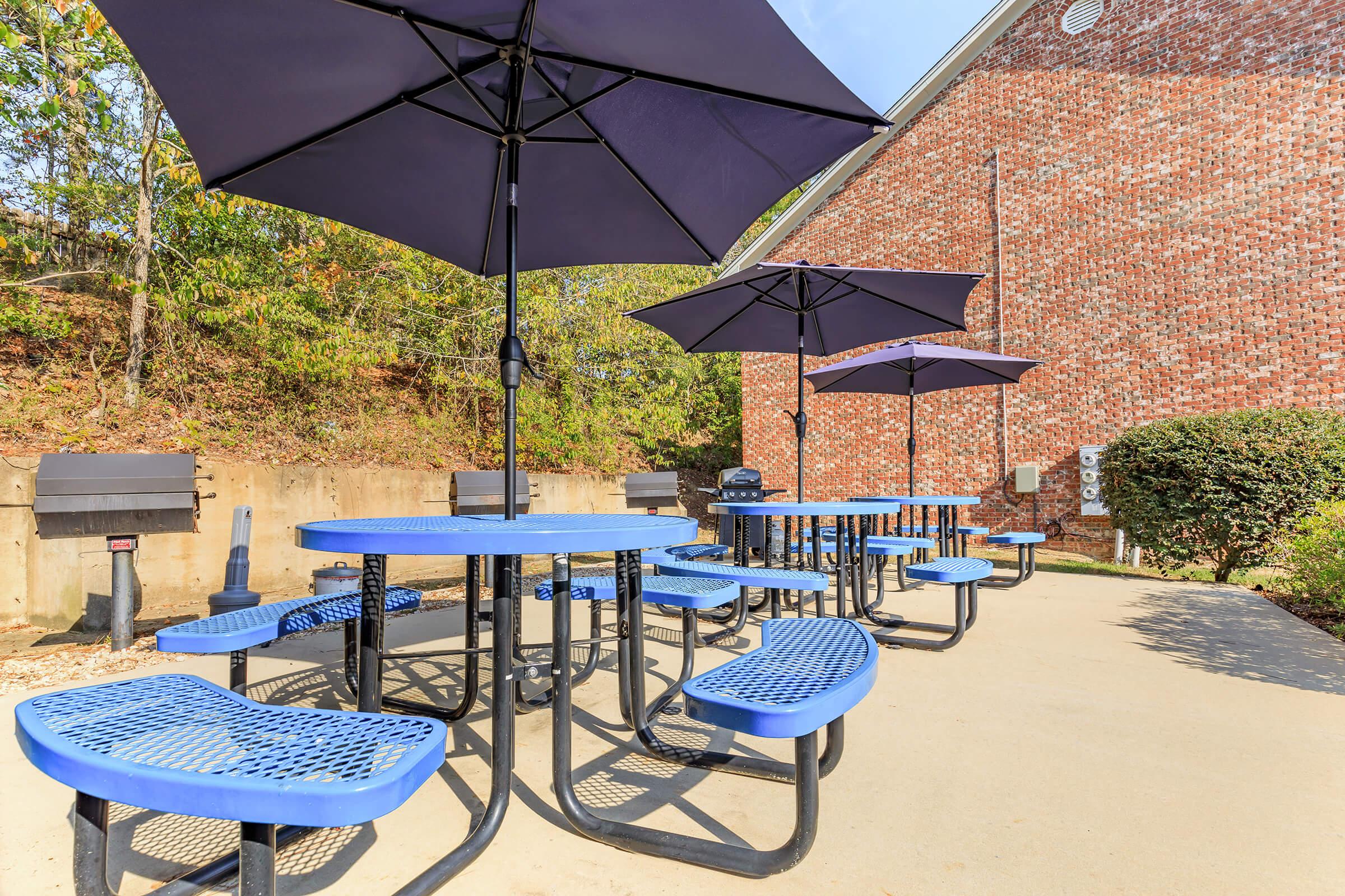 Blue picnic tables with umbrellas are set up in an outdoor area beside a brick building, surrounded by greenery. The scene is sunny, with a clear blue sky. There are multiple tables, each with matching blue chairs, providing a communal space for relaxation or dining.