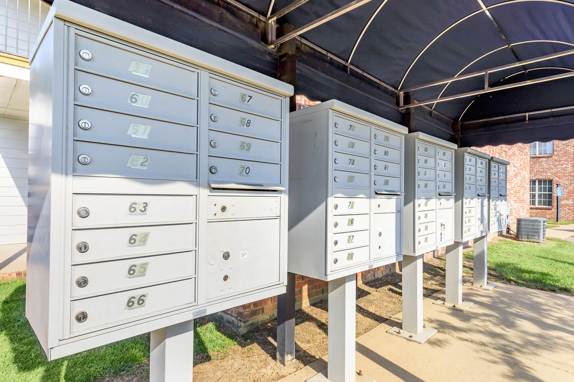 A row of mailbox units under a canopy, featuring multiple compartments numbered from 61 to 70. The mailboxes are metallic gray and positioned on pedestals. Lush green grass is visible around the mailbox area, with a brick building and an air conditioning unit in the background.