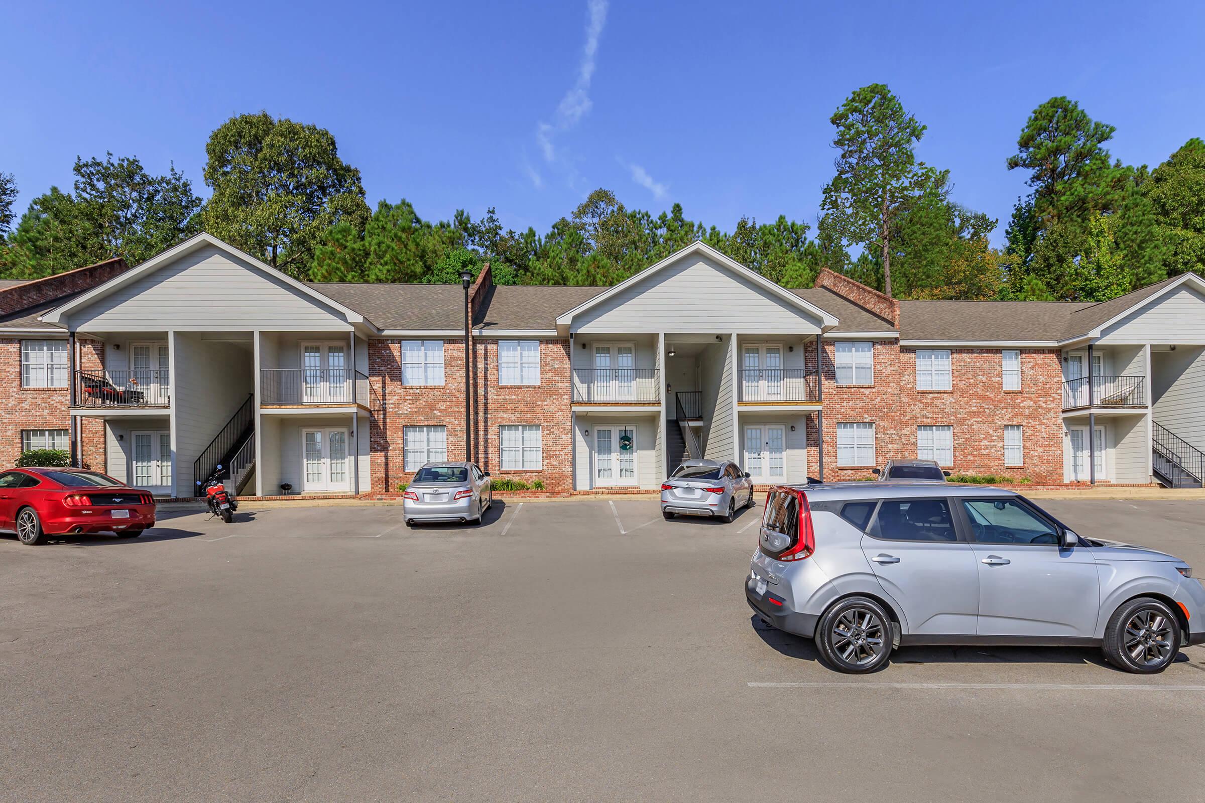 A view of a two-story apartment complex with a brick facade. Several parked cars are visible in the foreground, including a red car, a silver car, and a motorcycle. The surrounding area features trees and a clear blue sky.