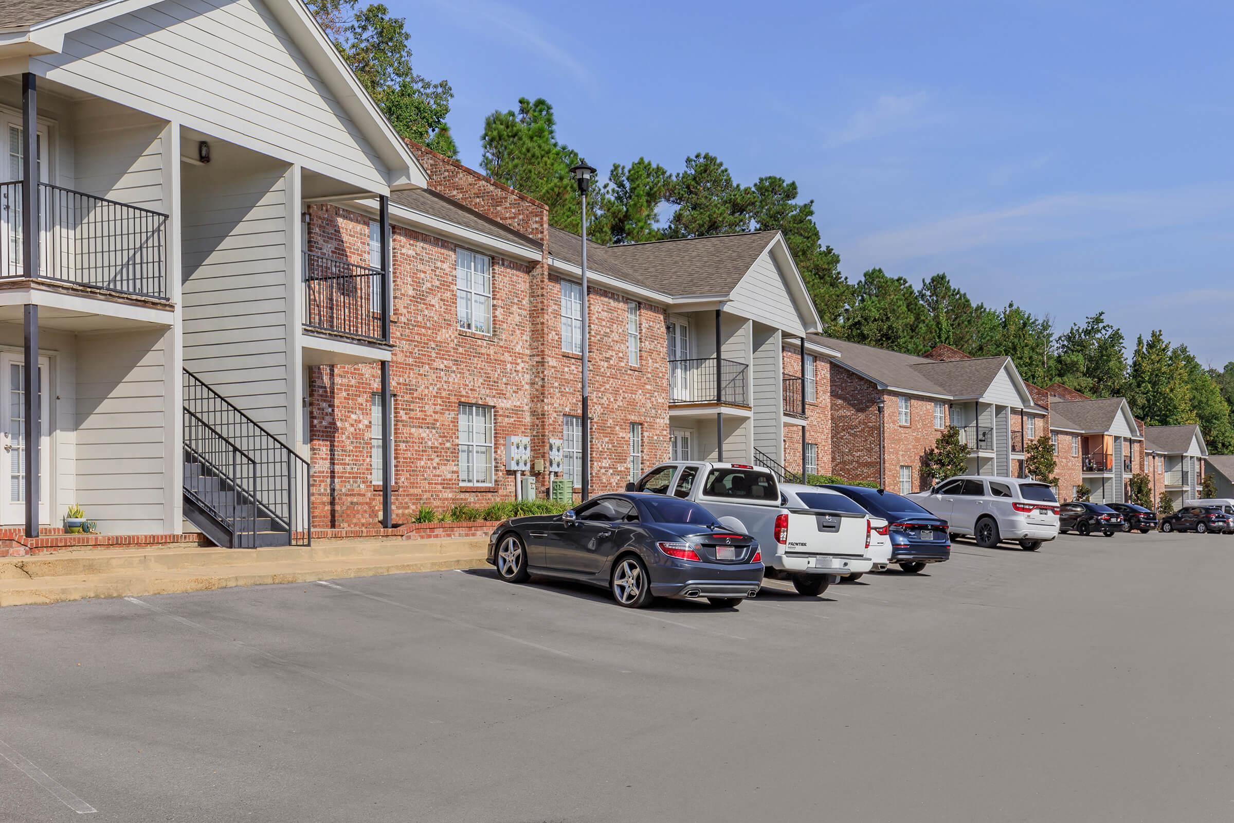 View of a residential apartment complex featuring two-story brick and siding buildings. A parking lot filled with various cars is visible in front of the apartments. The setting is surrounded by trees and blue skies, suggesting a pleasant, well-maintained area.