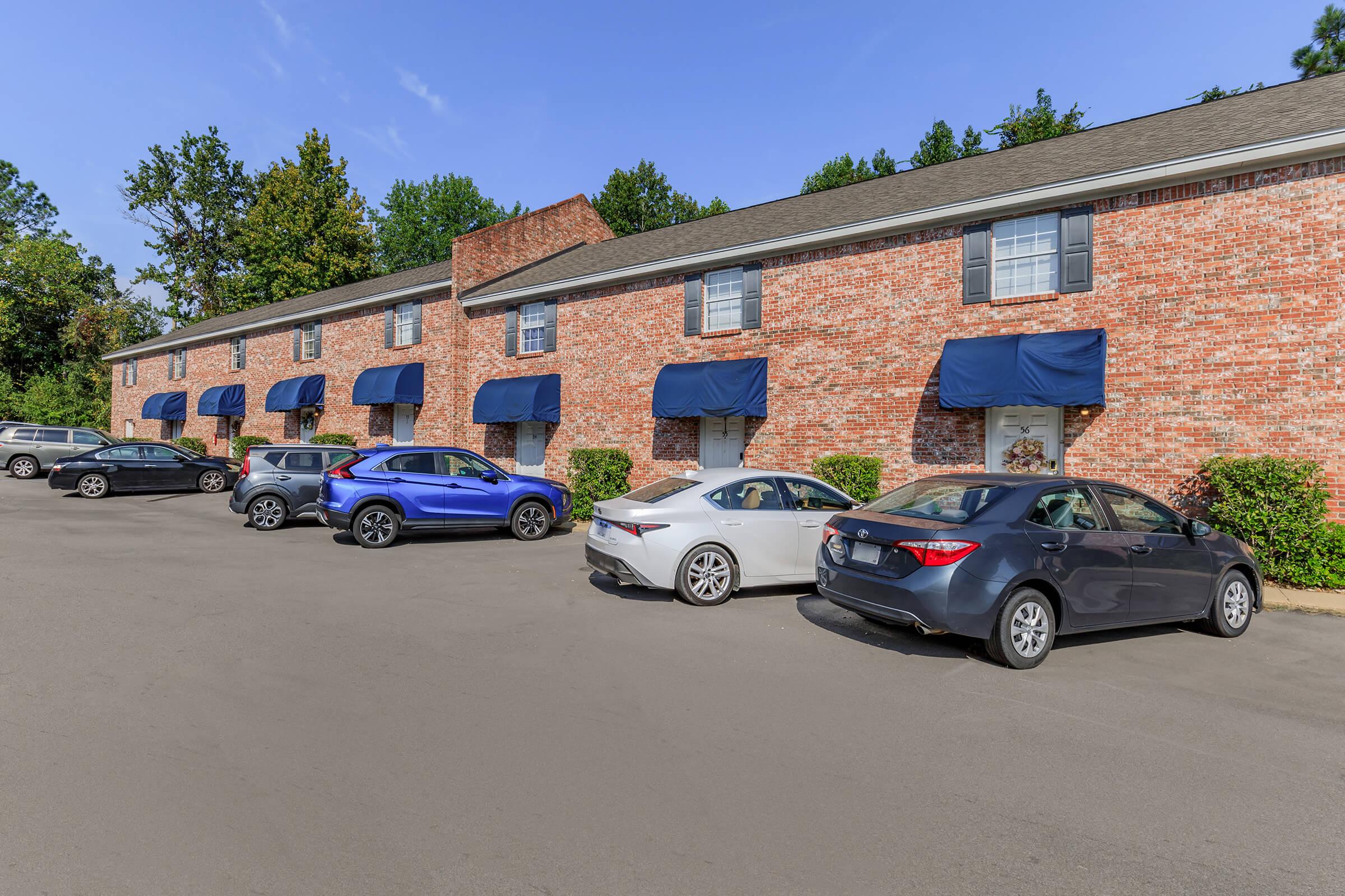 Row of brick buildings with blue awnings, featuring multiple parked cars, including a blue SUV, a silver sedan, and a gray vehicle. The setting has trees in the background under a clear blue sky.
