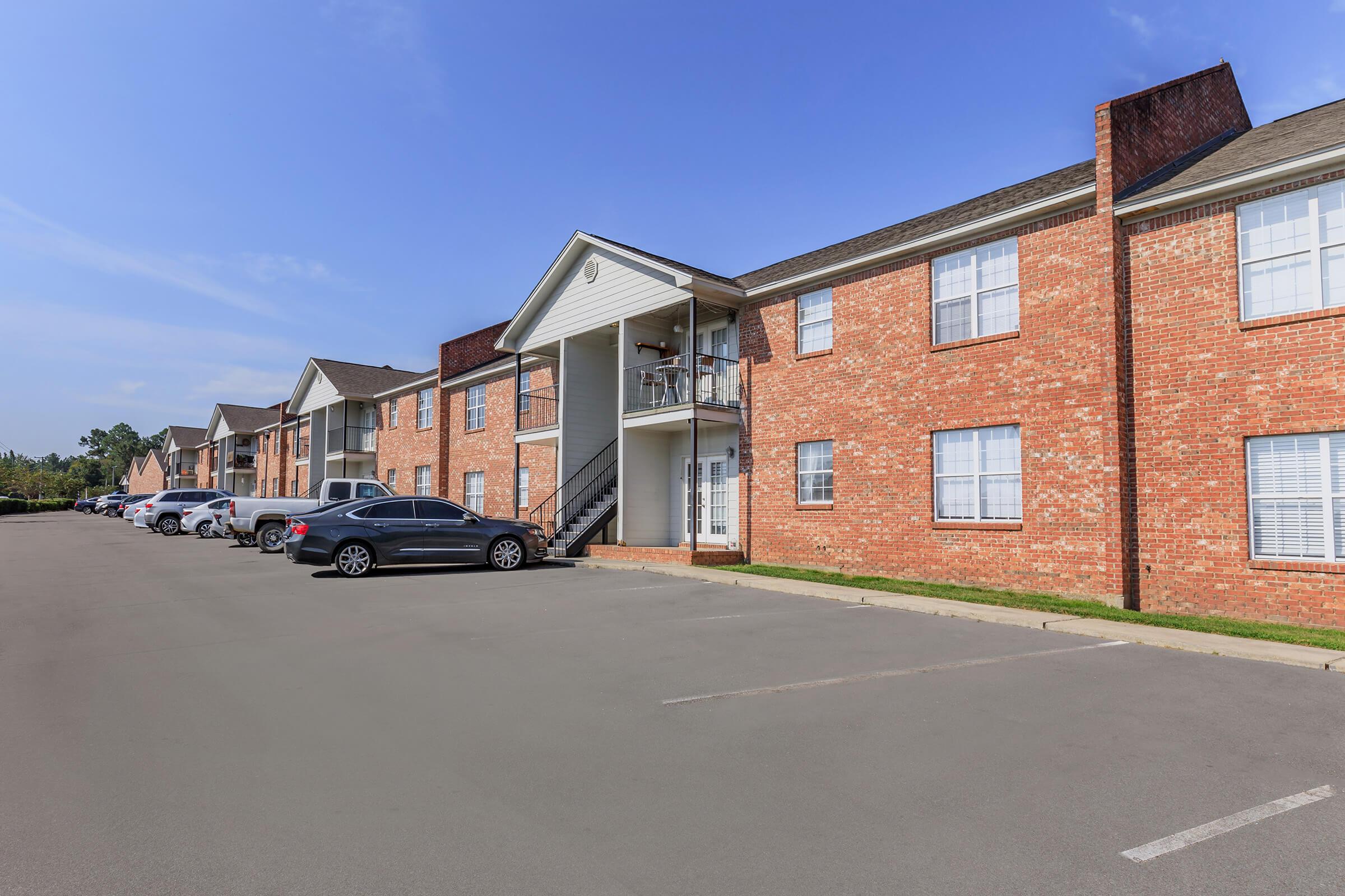 A multi-unit brick apartment building with two stories, featuring several parked cars in the foreground. The building has a set of stairs leading to the second floor balcony and is surrounded by green grass under a clear blue sky.