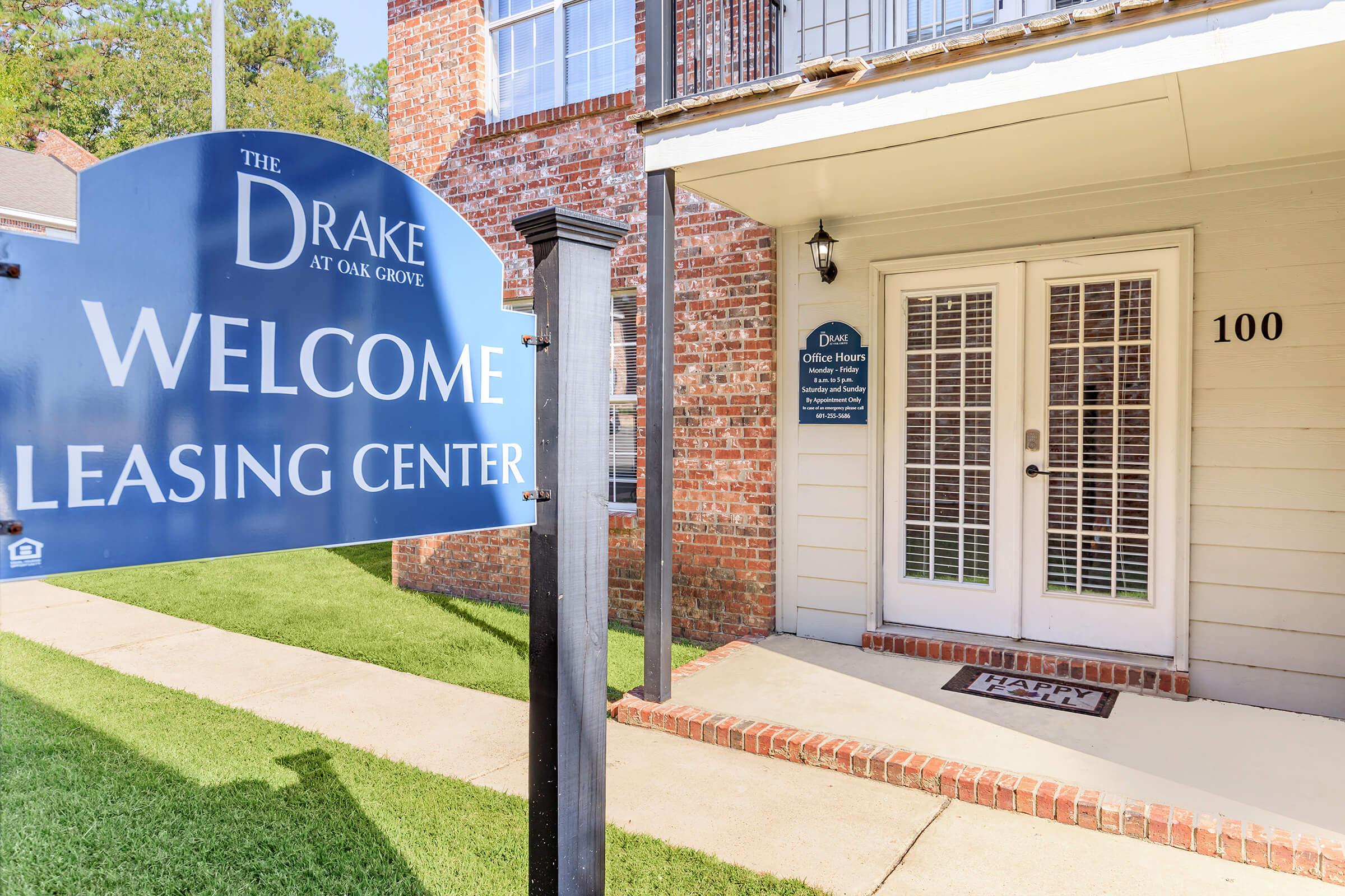Signage for "The Drake at Oak Grove" leasing center, featuring a prominent "Welcome" message. The entrance includes double doors with glass panels, and a small welcome mat is placed at the entrance. The building has a brick exterior and well-maintained lawn space.