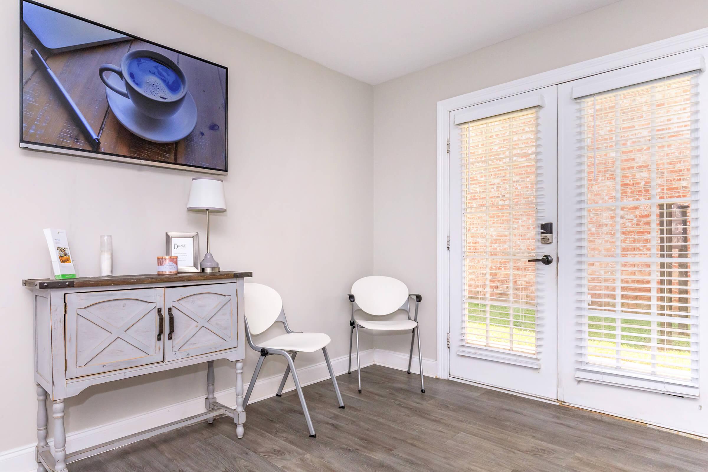 A modern waiting area features two white chairs, a table with decorative items, and a television mounted on the wall displaying an image of a coffee cup. Natural light streams in through glass doors with blinds, leading to an outdoor space. The walls are light-colored, creating a bright atmosphere.