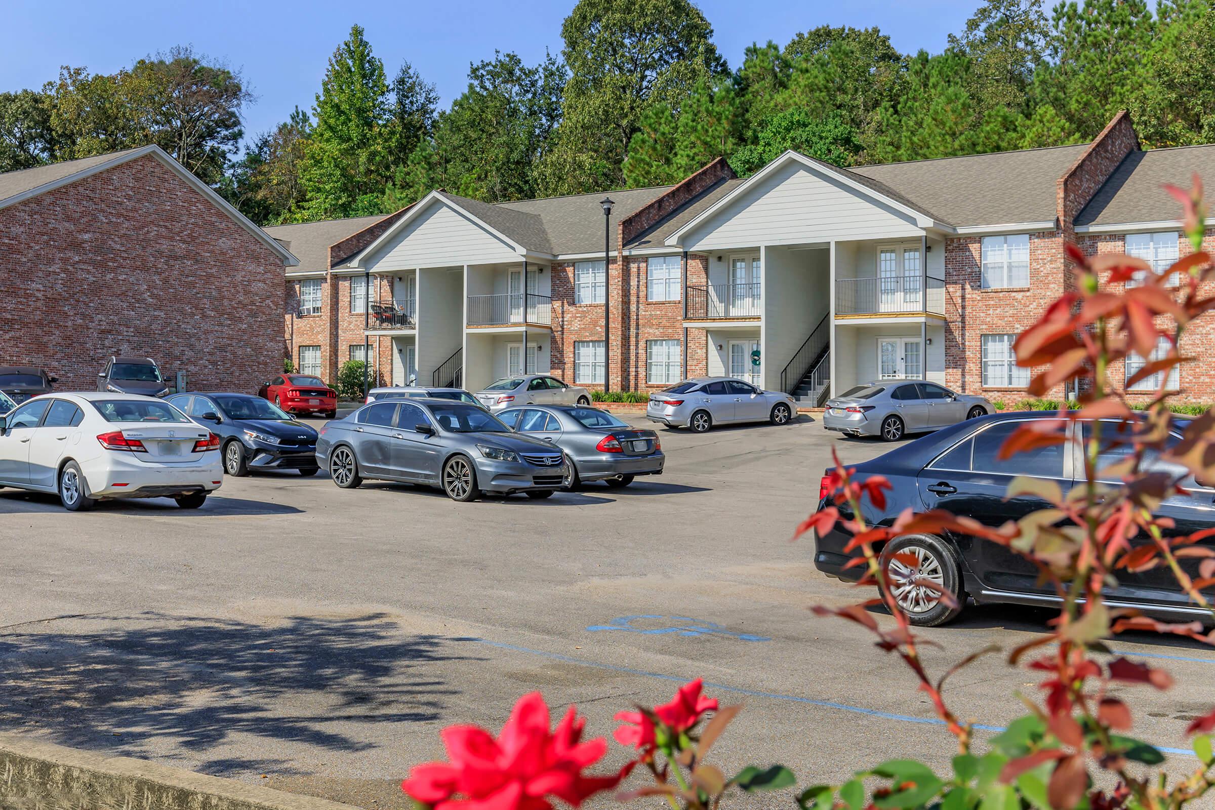 A parking lot in front of a two-story brick apartment building. Several cars of different makes and colors are parked in the lot. In the foreground, some red flowers and green foliage add color to the scene, with trees visible in the background. The sky is clear and sunny.