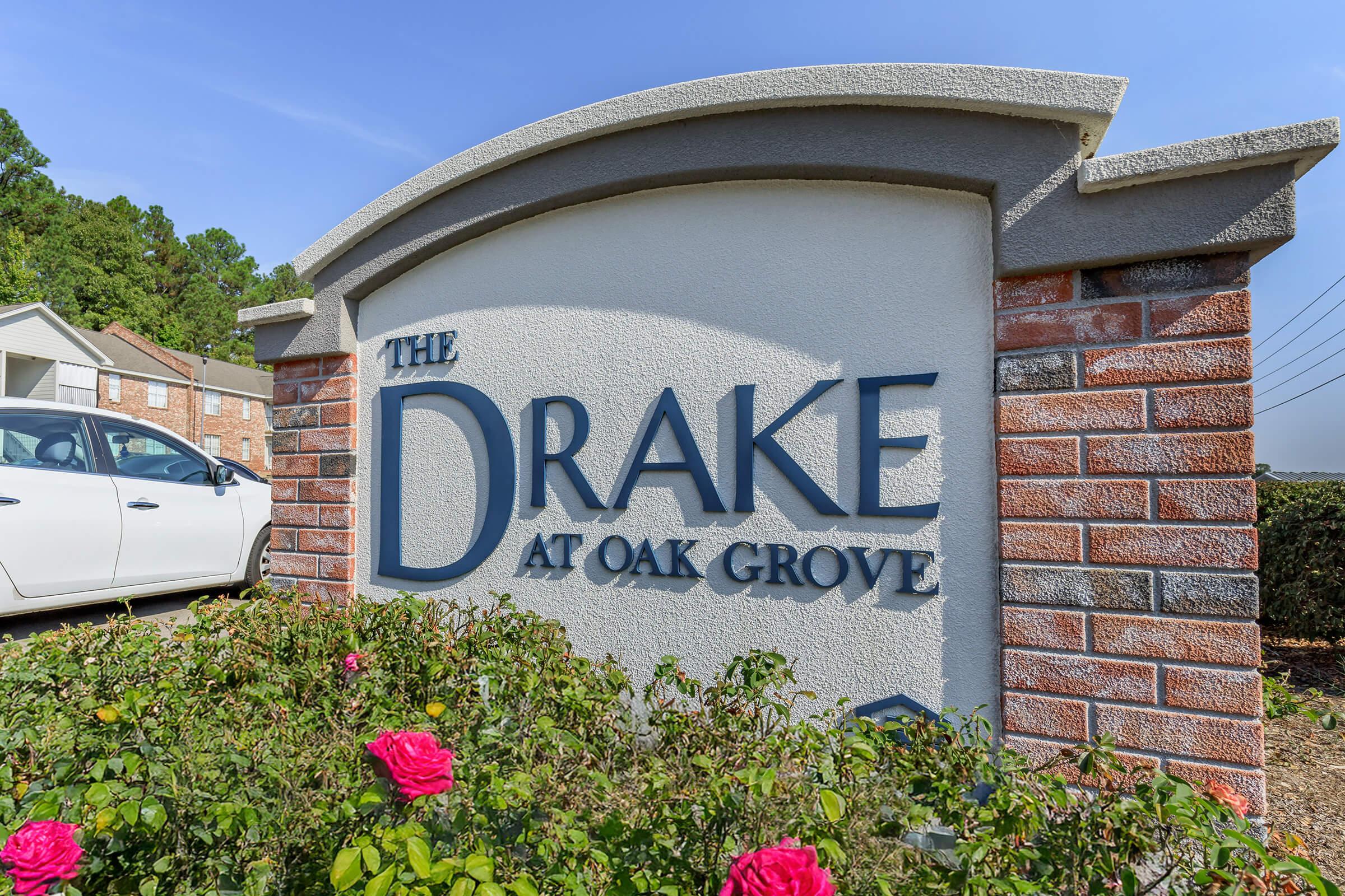 Sign for "The Drake at Oak Grove" featuring bold lettering on a decorative background. The sign is flanked by colorful rose bushes and set against a clear blue sky. In the background, there are buildings and power lines, suggesting a residential area.