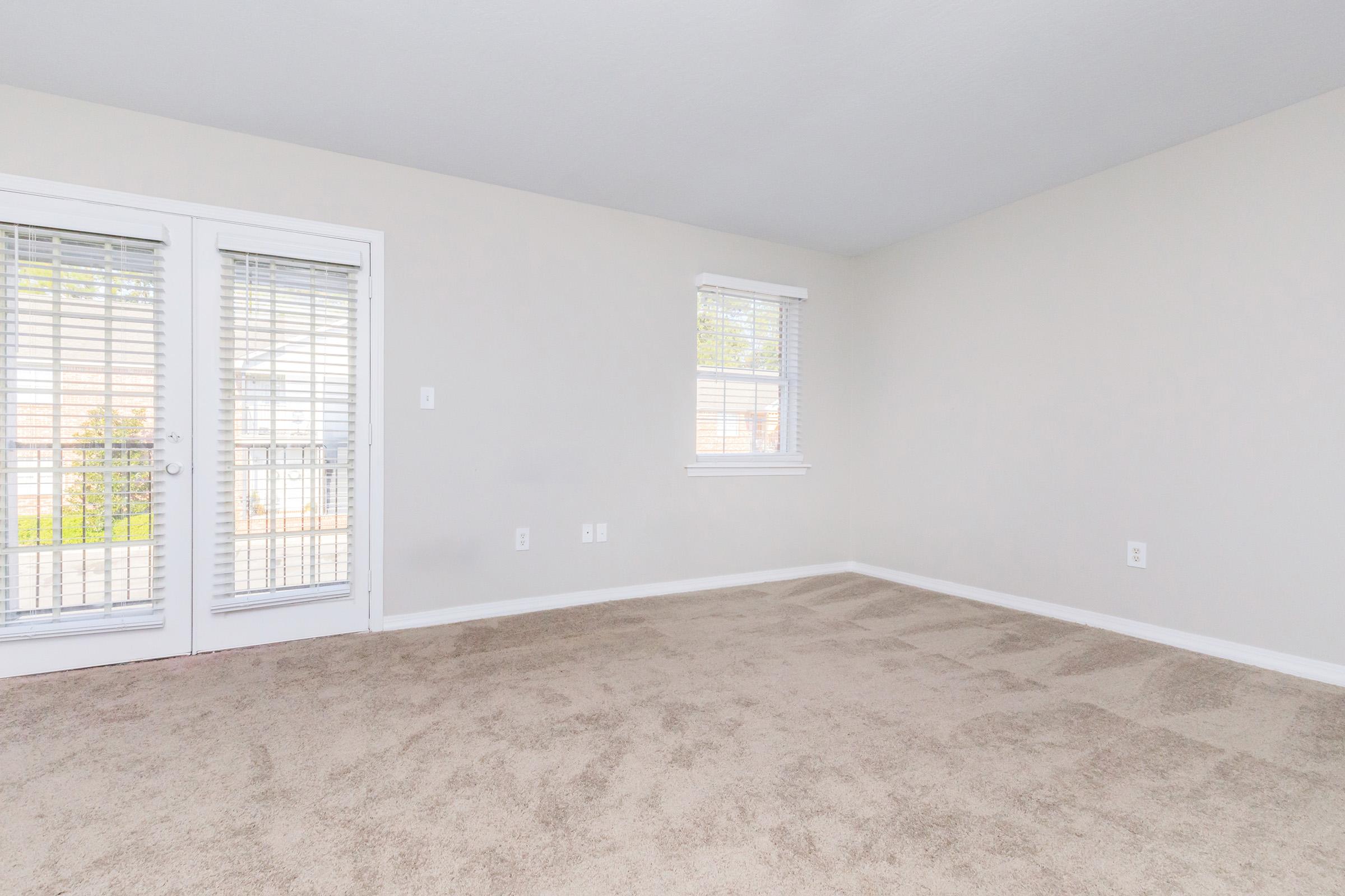 A light-filled, empty room with beige walls and plush carpet. There are two windows, one featuring vertical blinds and another set of glass doors leading to a balcony. The space appears clean and ready for furnishing, providing a neutral backdrop for various interior design options.