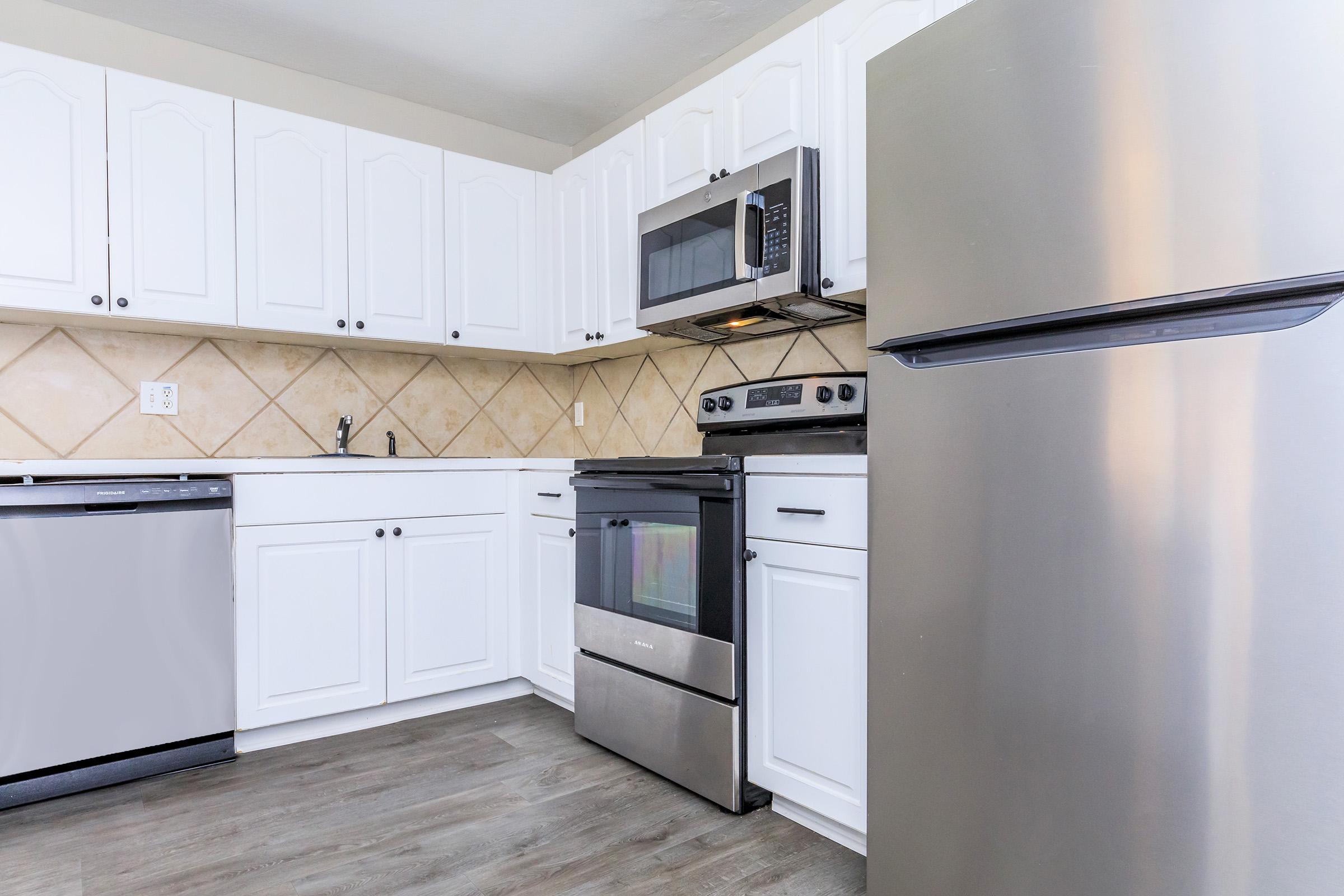 A modern kitchen featuring white cabinets, stainless steel appliances including a microwave and refrigerator, and a black oven. The countertops are light, with a tiled backsplash. The flooring is a wood-like laminate, creating a clean and contemporary look.