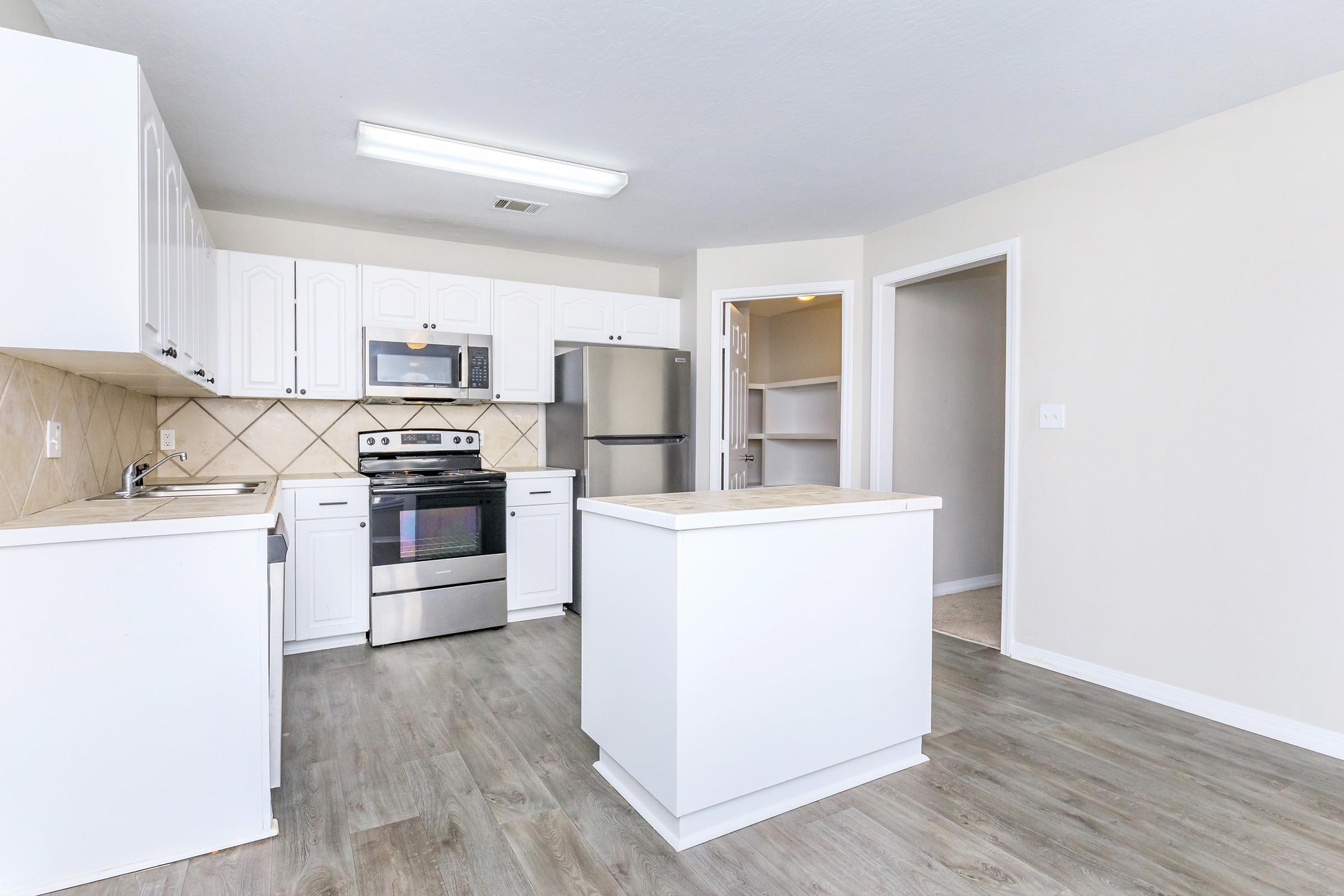 Modern kitchen with white cabinetry, stainless steel appliances including an oven and refrigerator, and a large kitchen island. The floor is light-colored laminate, and there is a spacious pantry accessible through a door on the right. Natural light fills the area through a window above the sink.