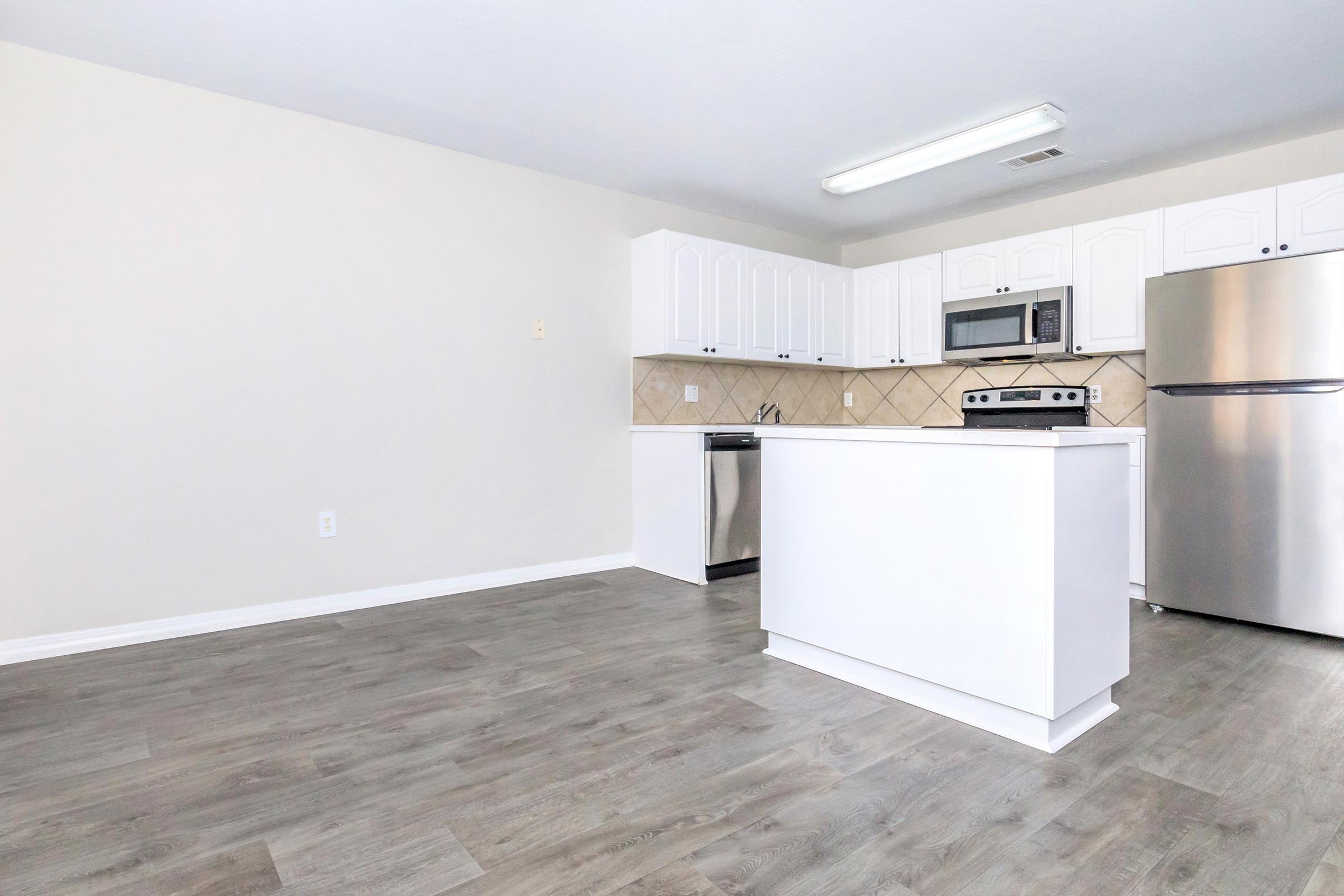 A modern kitchen featuring white cabinetry, stainless steel appliances, and a spacious layout with a light wood floor. The area is well-lit with overhead lighting and has a neutral wall color, creating a clean and inviting atmosphere.