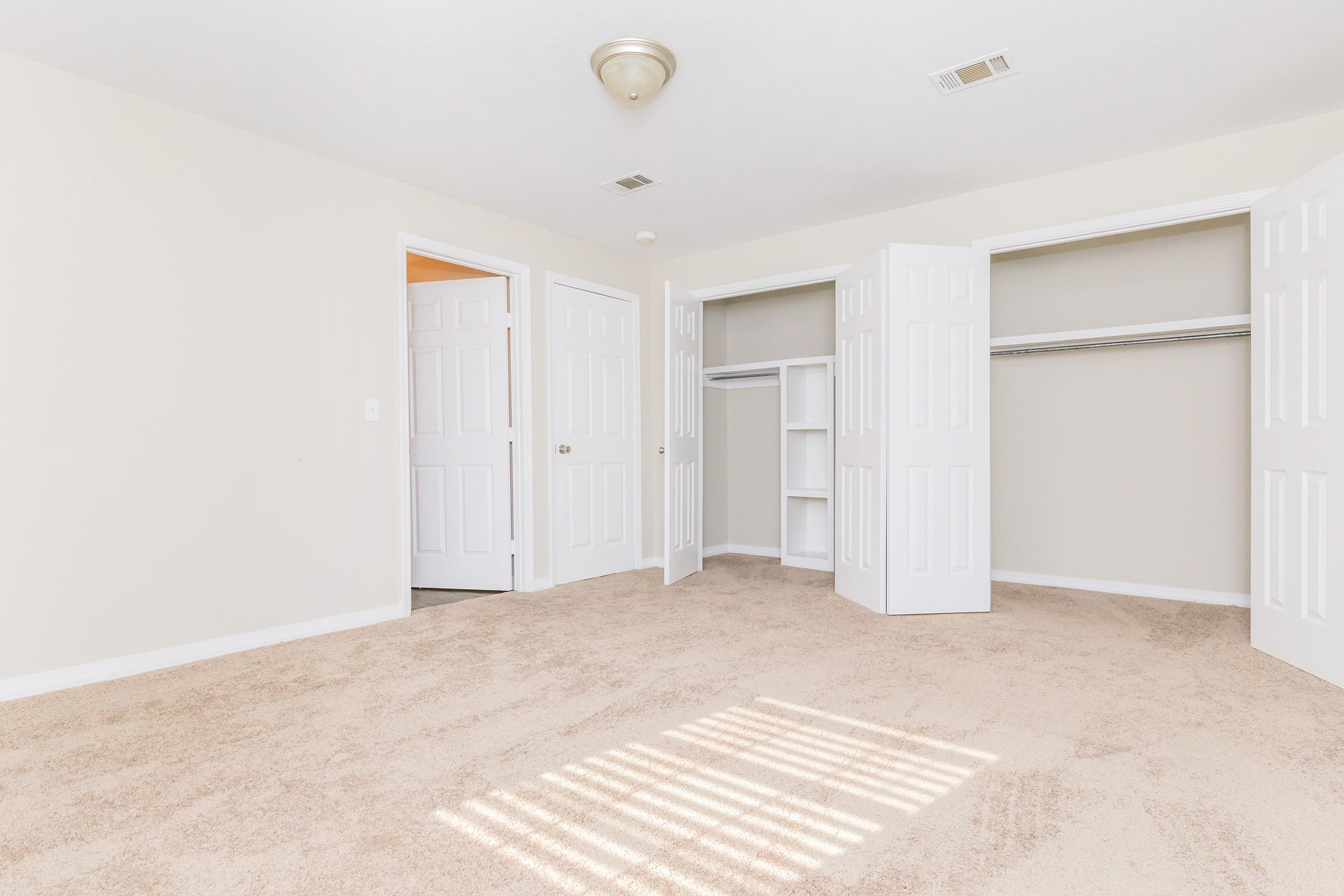 Empty bedroom featuring light-colored walls and plush beige carpeting. Two open closet doors reveal ample storage space. Natural light streams in through a window, casting shadows on the floor. Additional doors lead to an adjacent room, enhancing the spacious feel of the area.