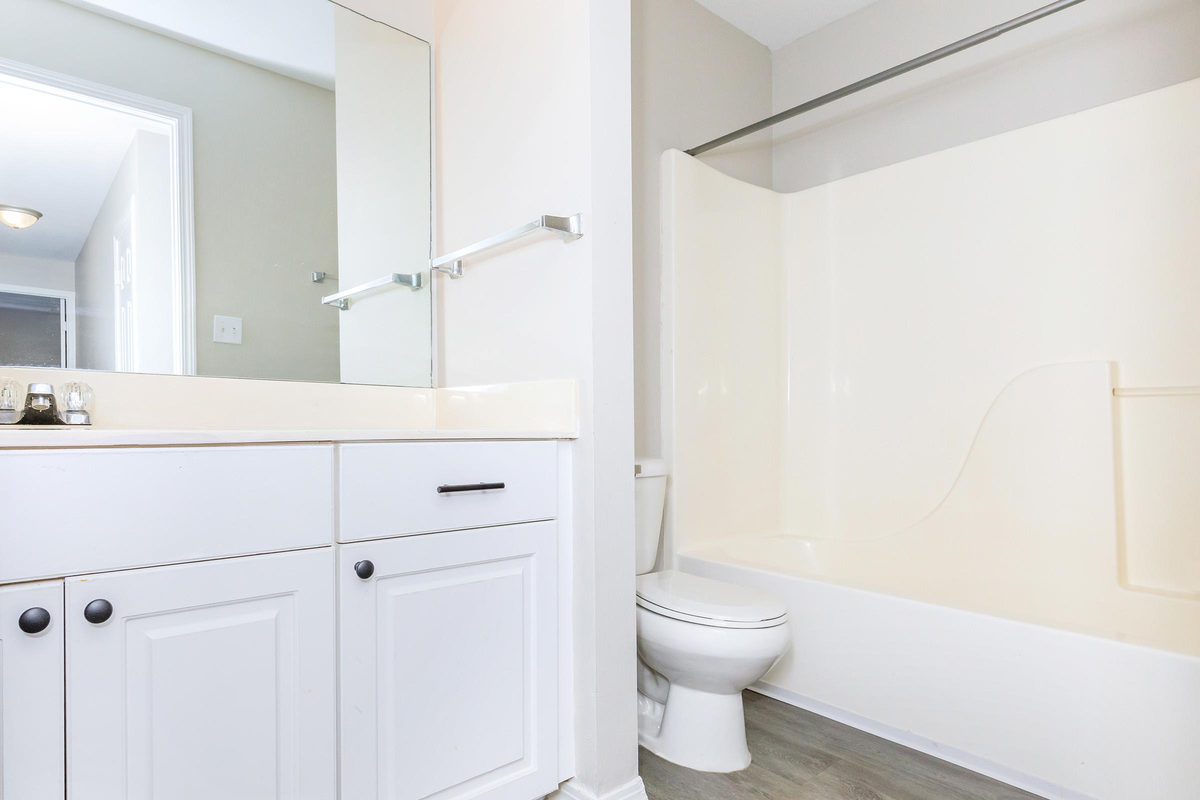 A clean, modern bathroom featuring a white bathtub, toilet, and a sink with white cabinets. The walls are light-colored, and there is a large mirror above the sink. The floor is light gray, and the overall appearance is bright and spacious.