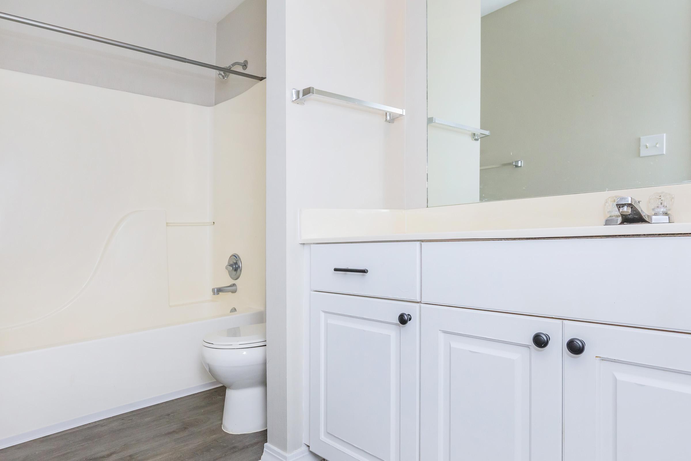 A clean and modern bathroom featuring a white bath tub, toilet, and a vanity with black knobs. The walls are painted in a light color, and there is a mirror above the sink. The floor has a wooden appearance, adding warmth to the space.